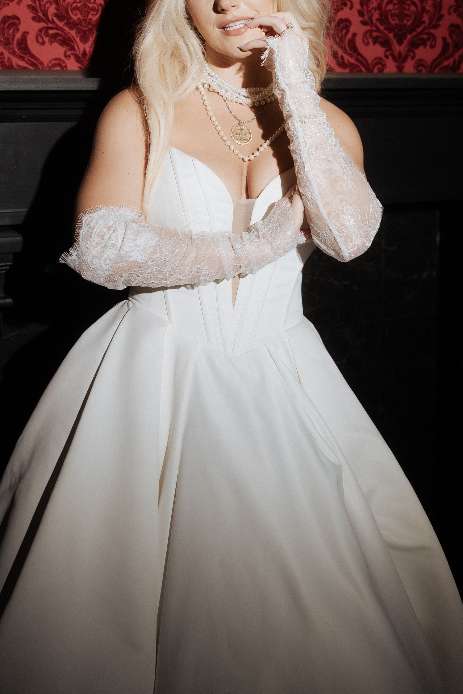 Elegant white wedding dress with lace sleeves and pearl necklace details photographed against dark red damask wallpaper.