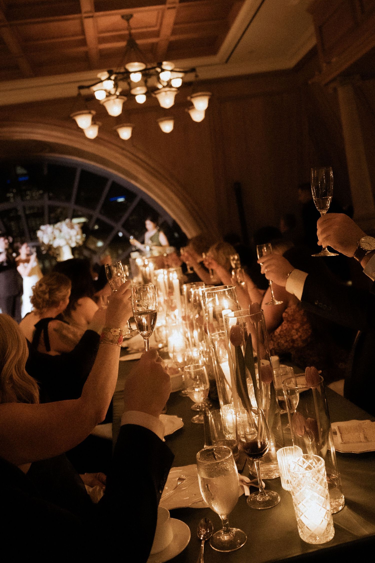 Elegant candlelit wedding reception dinner with guests raising champagne glasses for a toast in a grand ballroom.