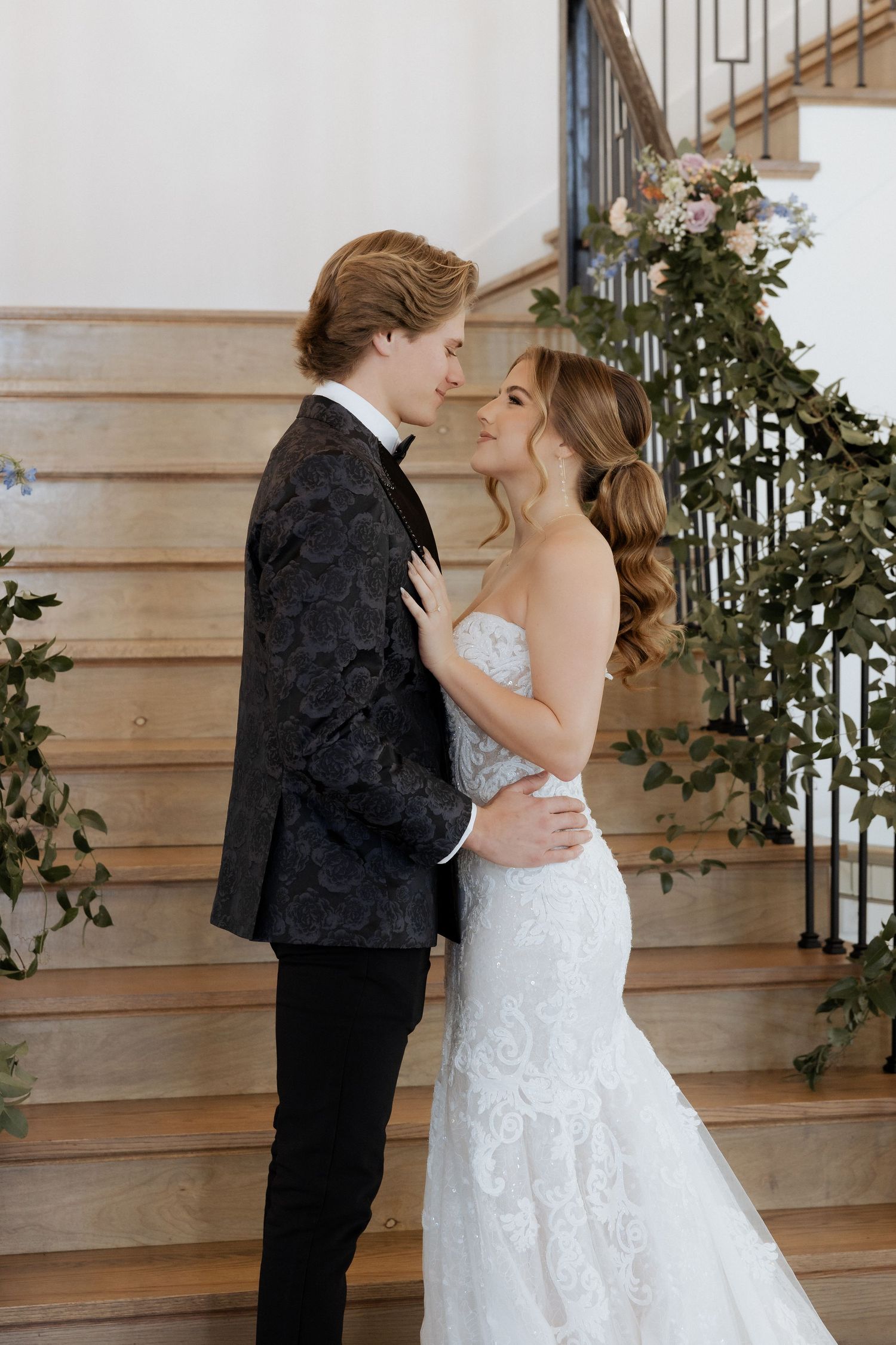 A romantic wedding photo of a couple embracing by a wooden staircase with decorative greenery.