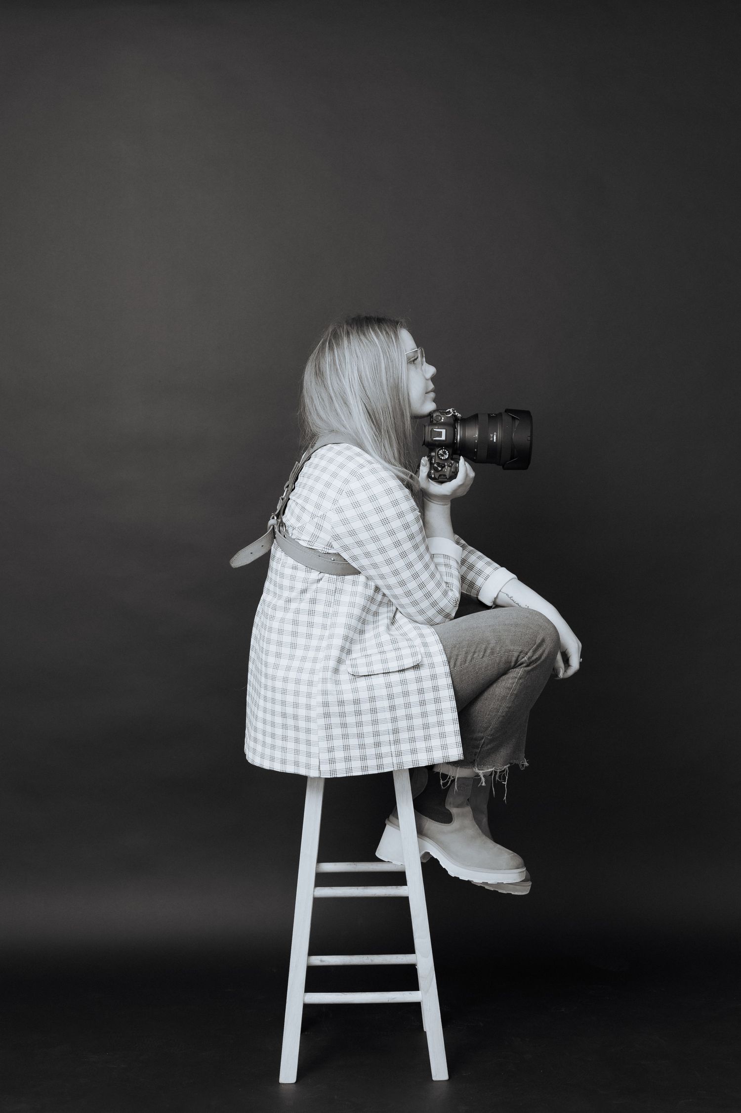 A photographer in a checkered dress perches on a white stool with a professional camera against a dark background.