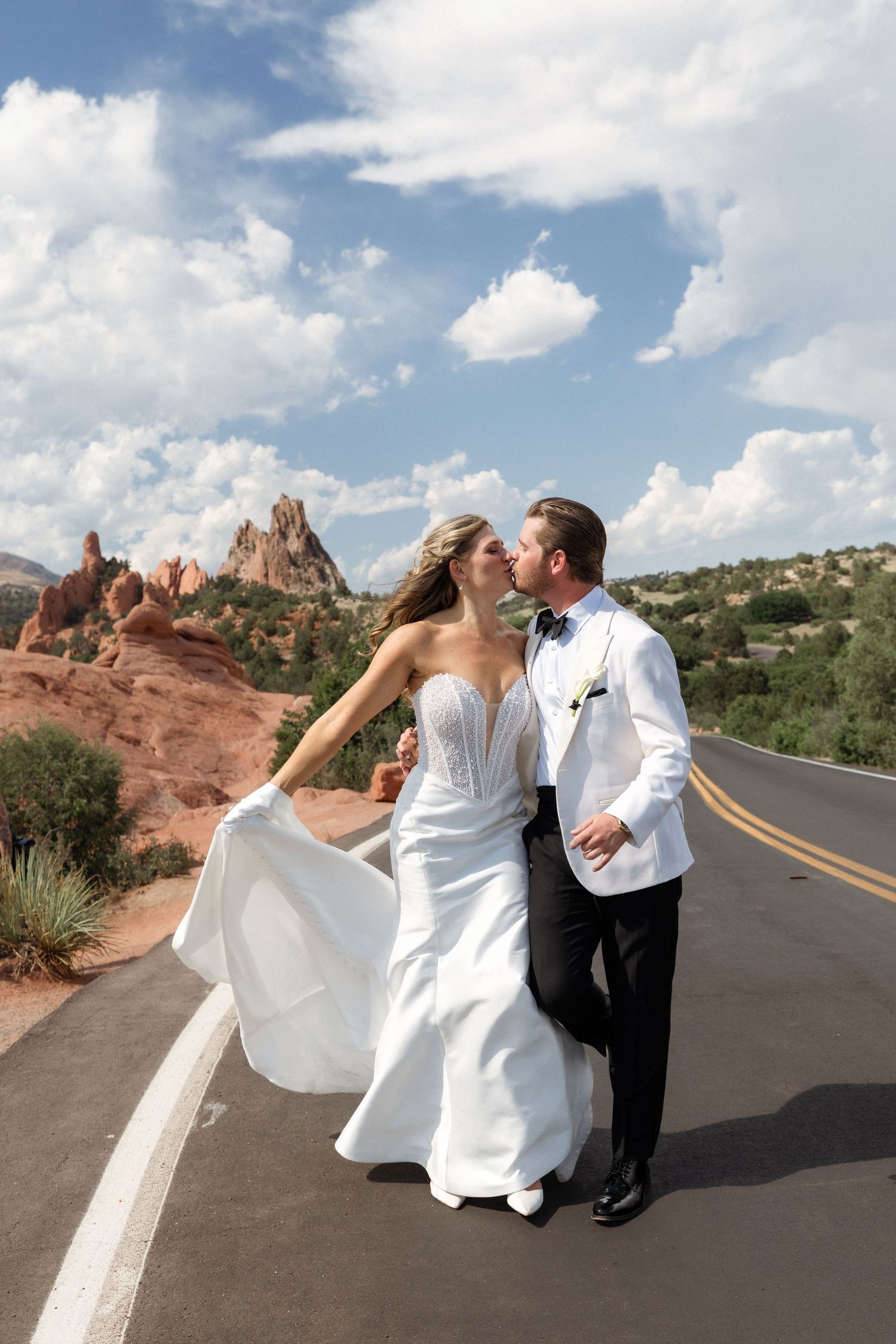 Bride and groom share a kiss on a scenic road at Garden of the Gods, Colorado, with red rock formations under a blue sky.