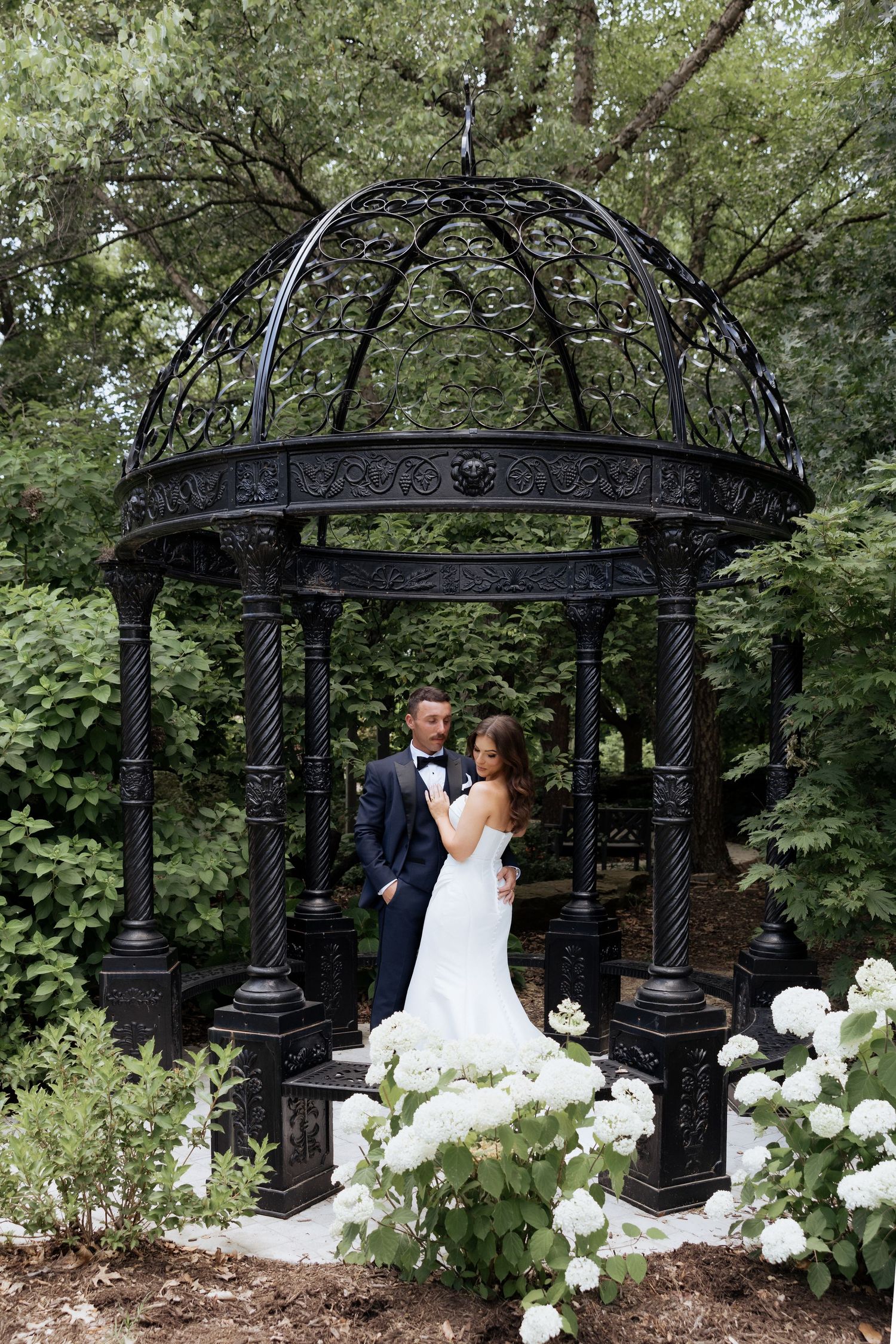 Bride and groom posing under an ornate black iron gazebo surrounded by lush greenery and white hydrangeas.