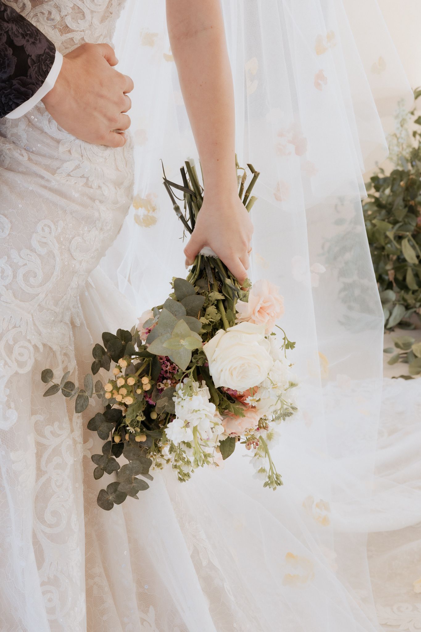 A delicate bridal bouquet with white roses and eucalyptus leaves held against a flowing lace wedding dress.