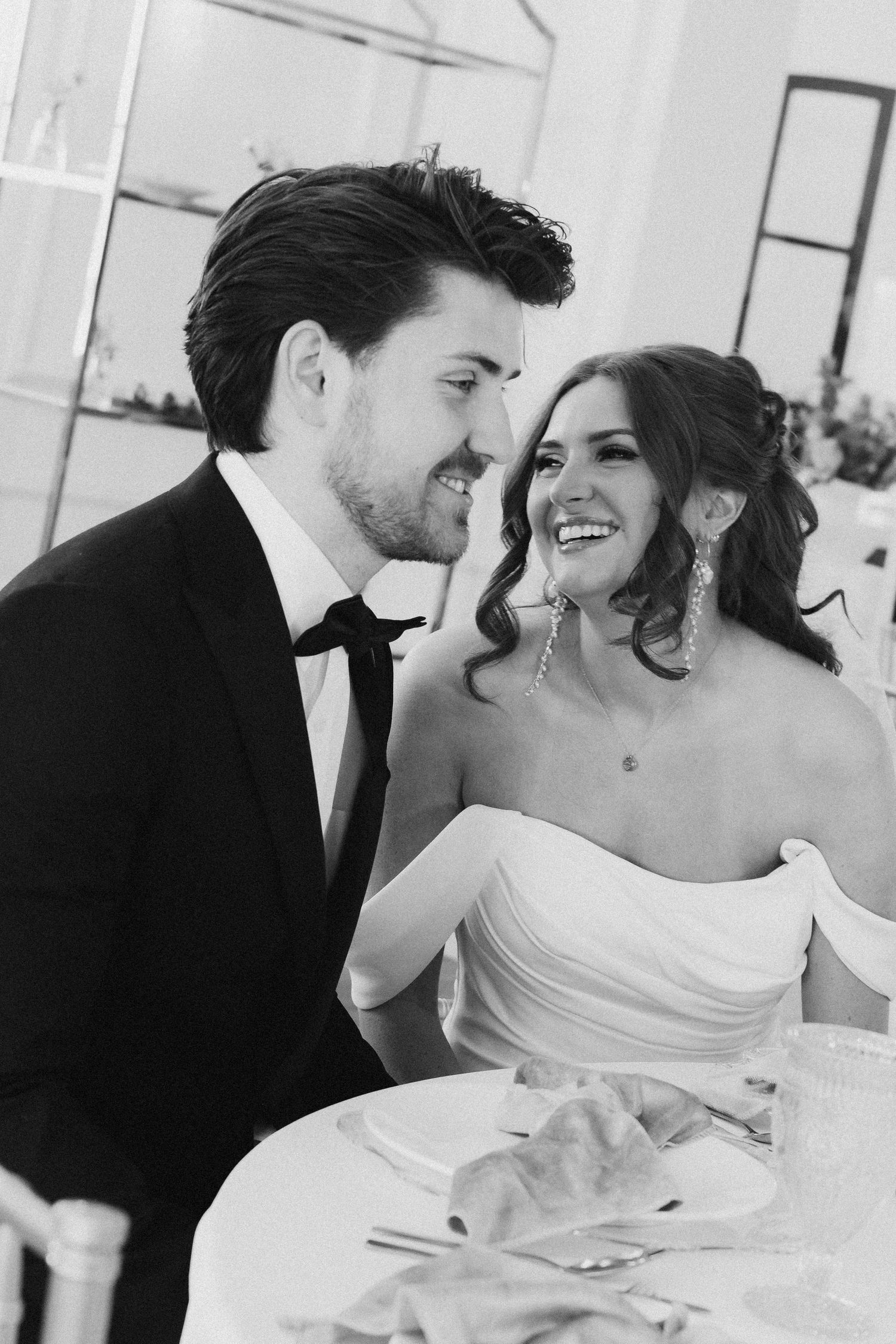 A joyful black and white candid photo of a bride in a strapless gown and groom in a tuxedo sharing a moment at their wedding.