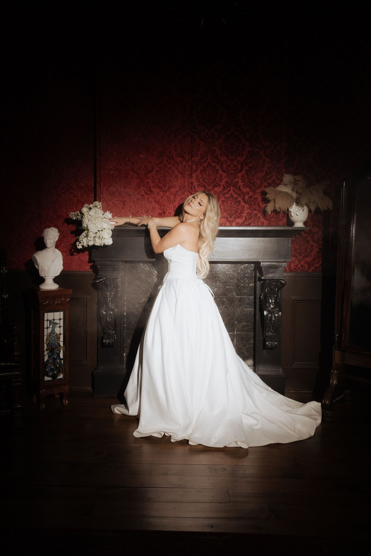 A bride in a flowing white wedding gown poses elegantly against a dark red damask wall with decorative white busts.