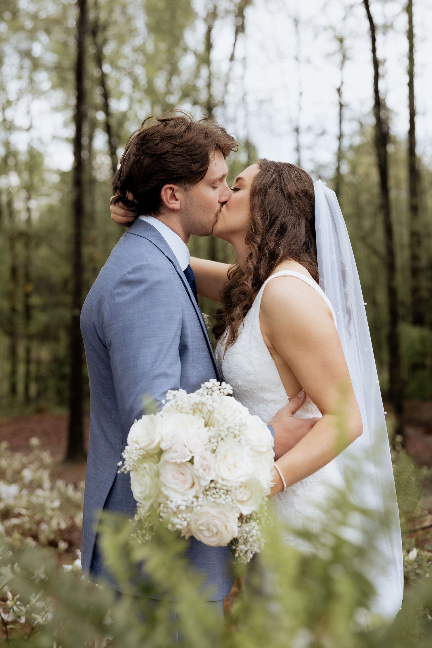 A wedding couple shares a romantic kiss in the forest while holding an elegant white floral bouquet.