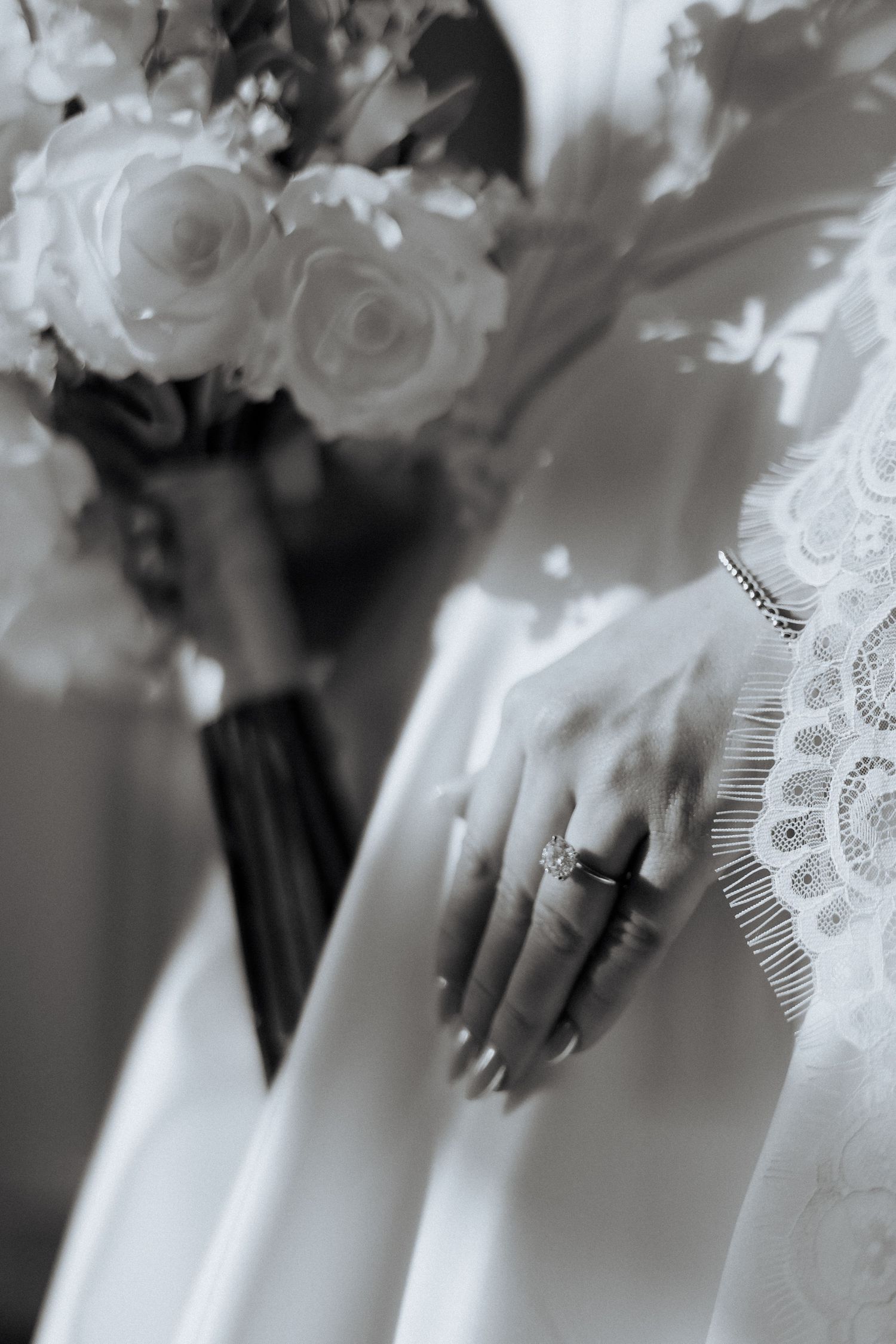 Close-up black and white photo of roses in bridal bouquet with lace details and engagement ring.