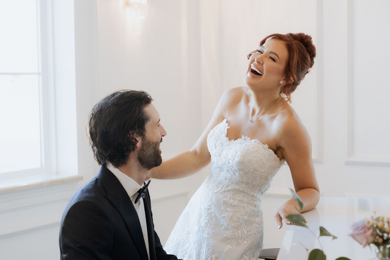 A joyful wedding moment as a bride in white dress shares a laugh with someone in a black suit near a window.