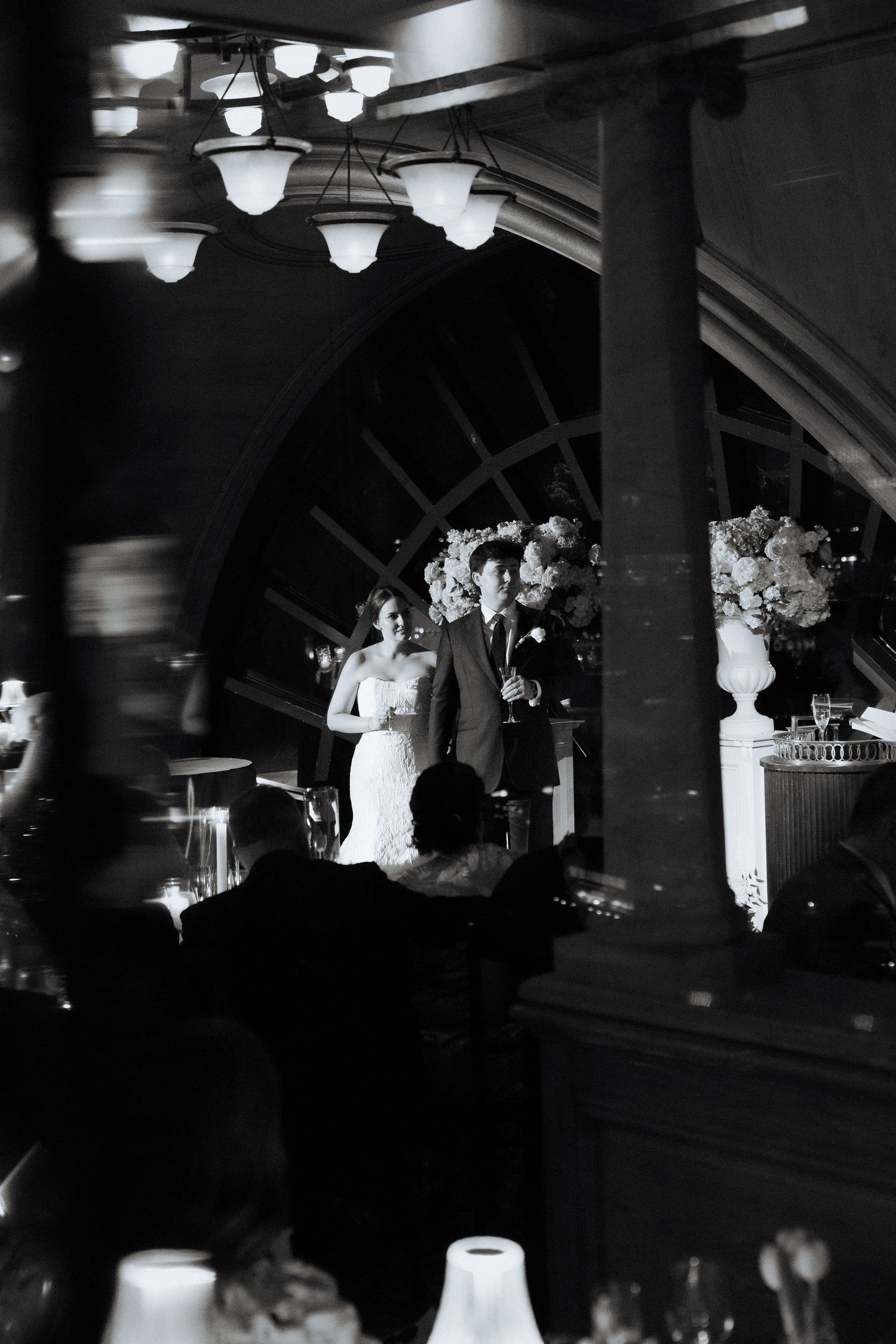 Black and white photo of a bride and groom standing together at their wedding reception beneath elegant chandeliers.
