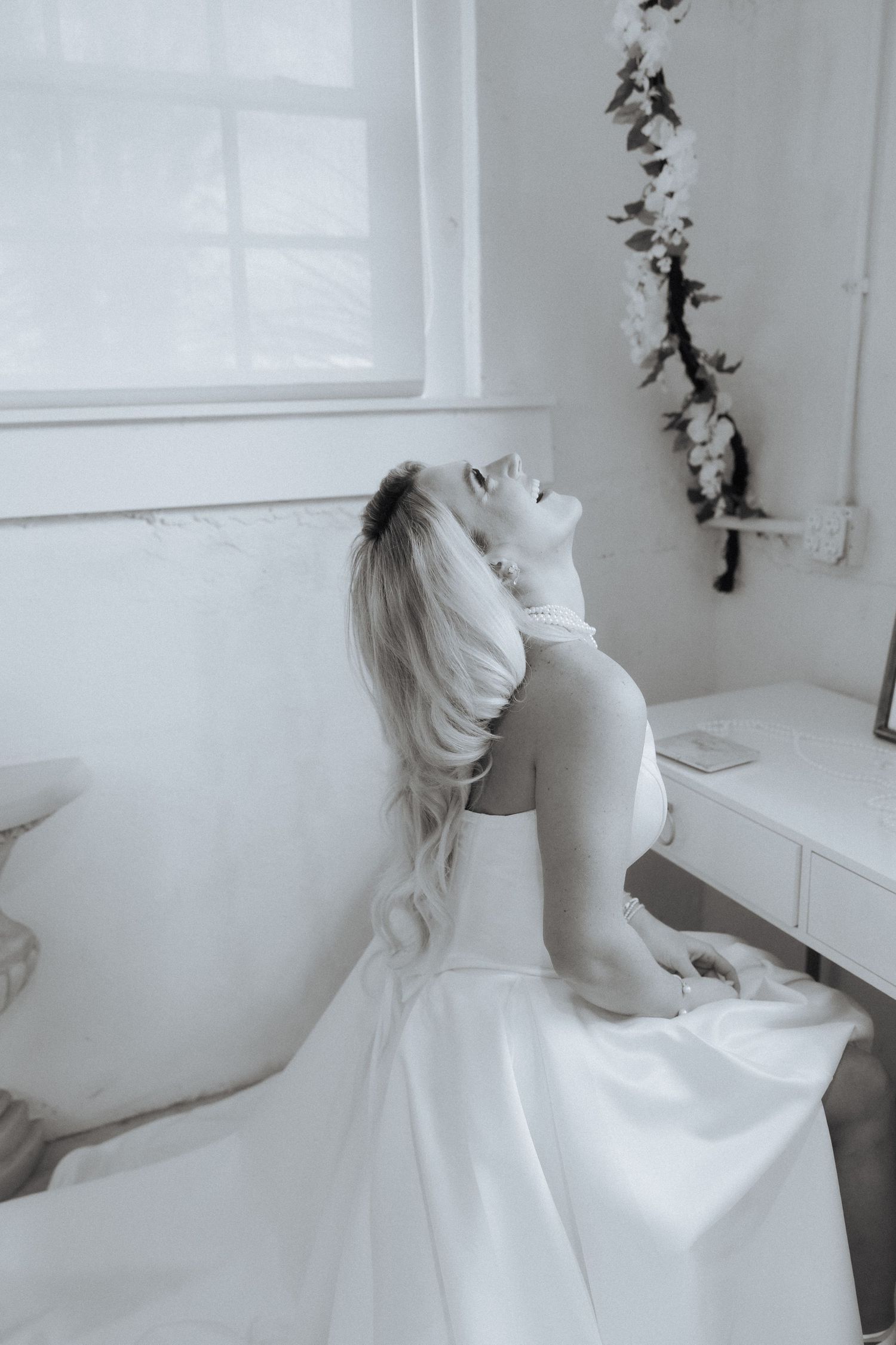 A figure in a white gown gazes upward in an ethereal monochrome portrait set against a minimalist white room.