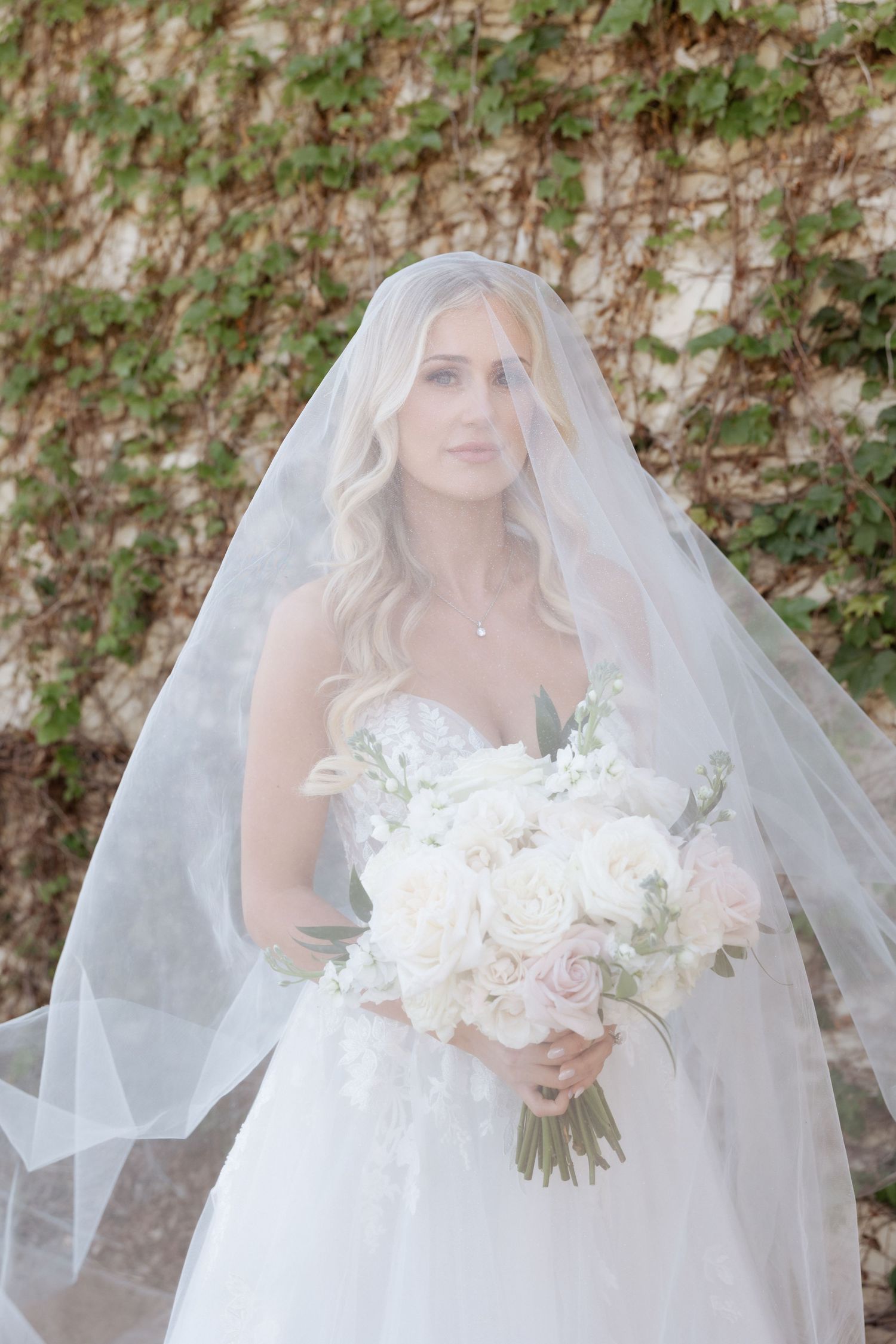 Bride in white lace gown and flowing veil holds white and blush rose bouquet outdoors near ivy-covered wall.