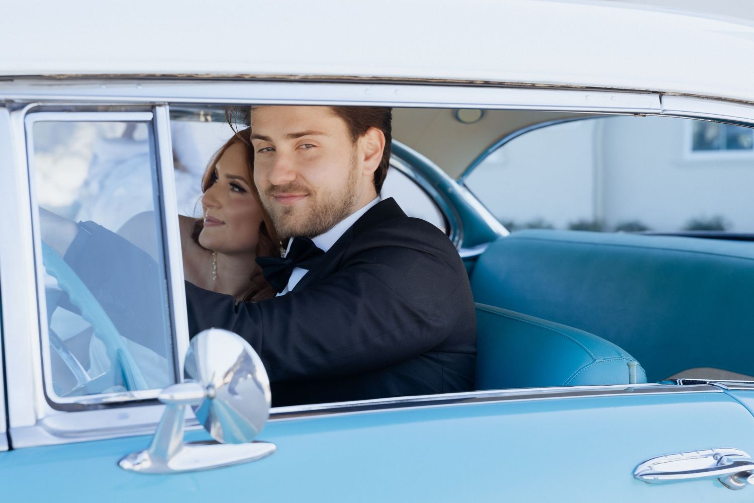 A couple sits in a vintage blue car dressed in formal attire.