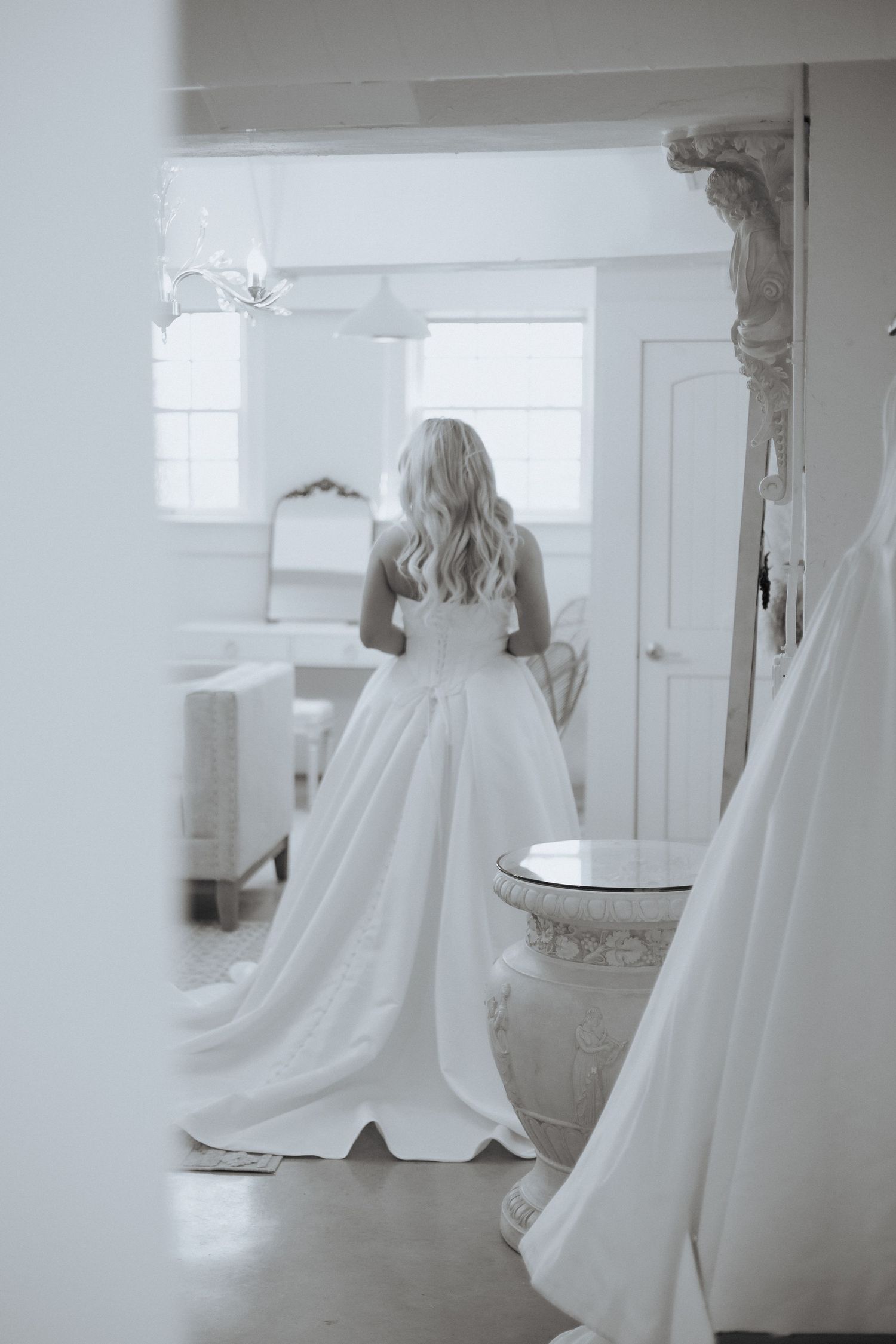 A bride in a flowing white wedding gown stands in an elegant bridal boutique with natural light streaming through windows.