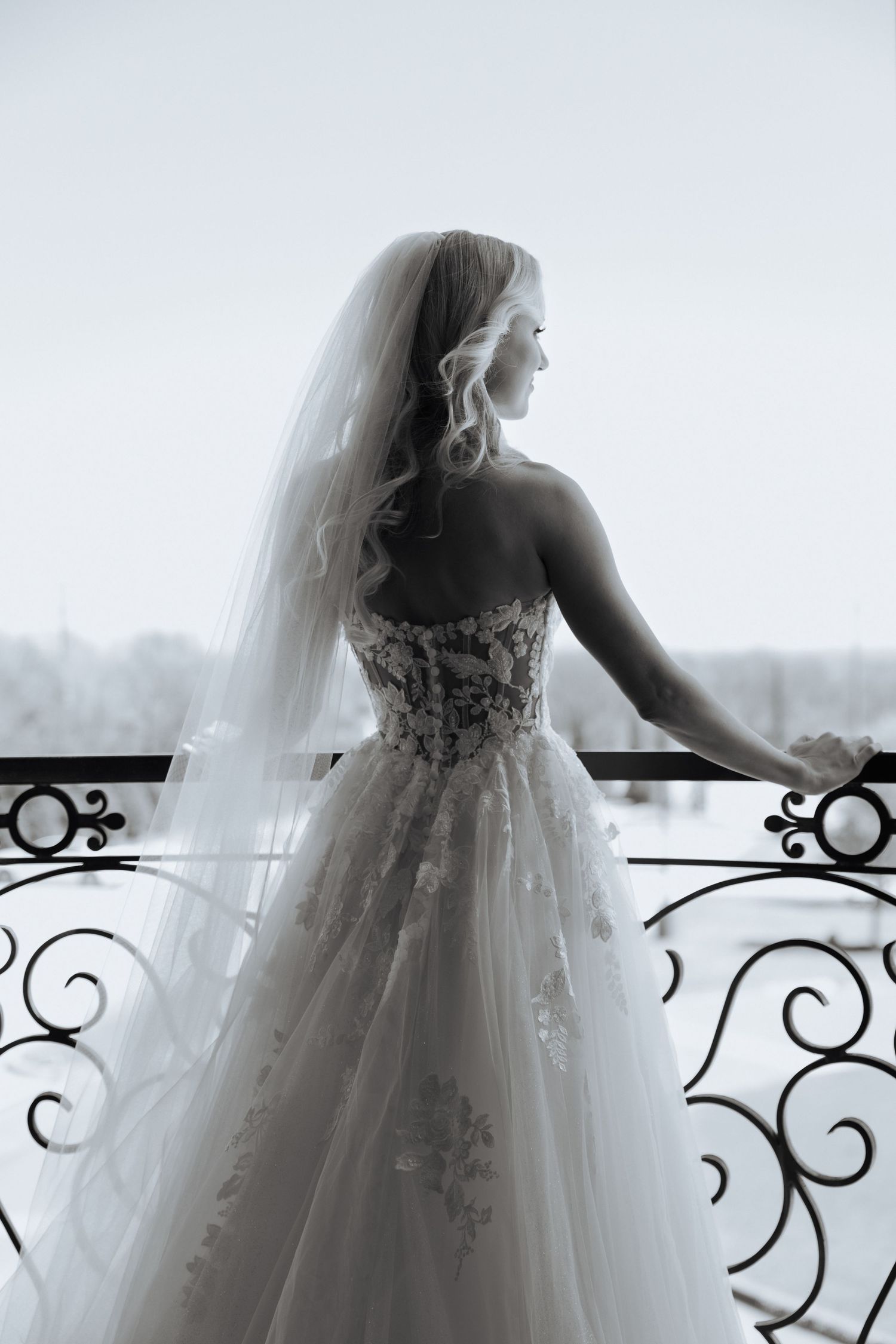 Elegant bride in a strapless lace ball gown and veil stands on an ornate balcony, gazing into a winter landscape.
