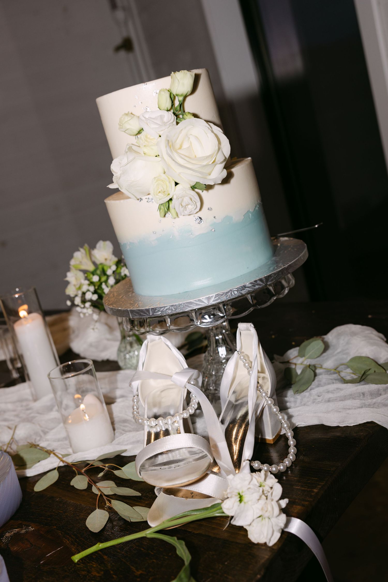 Elegant white wedding cake with light blue ombre design displayed on crystal cake stand with white floral decorations.
