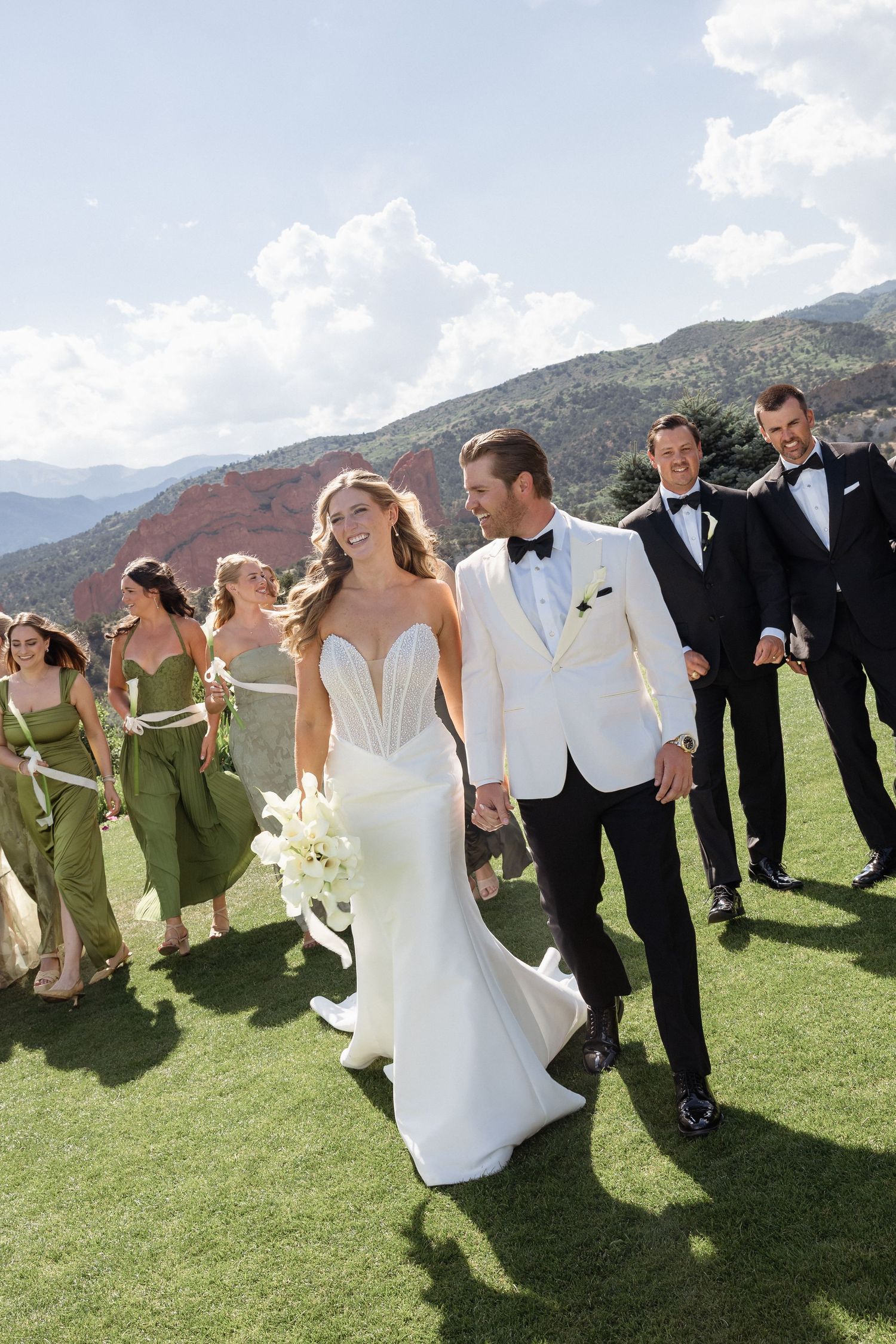 Bride and groom walk with wedding party in green dresses and black tuxedos against a scenic mountain backdrop.