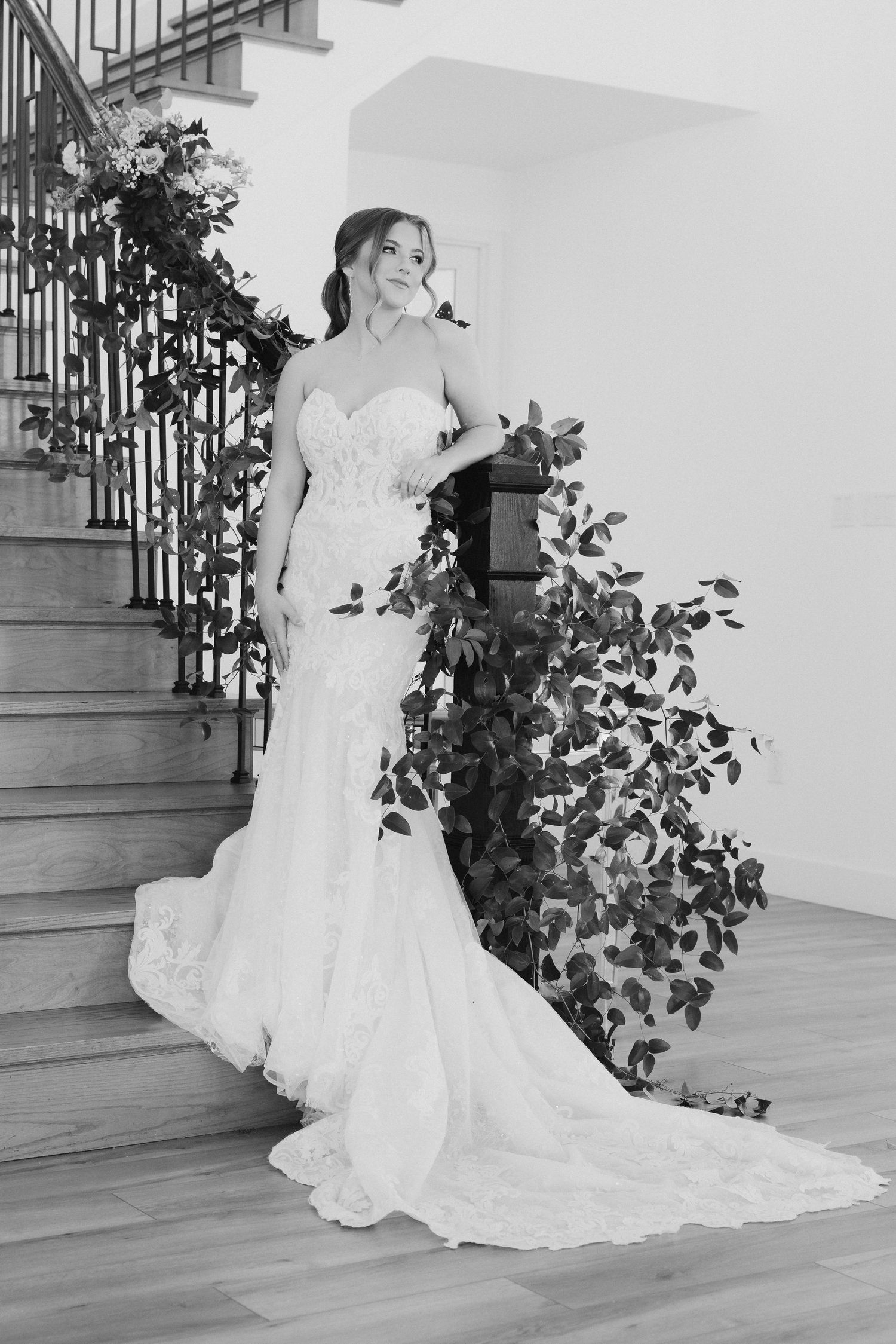 A bride in a fitted lace mermaid wedding gown with a flowing train poses elegantly by a staircase with greenery.