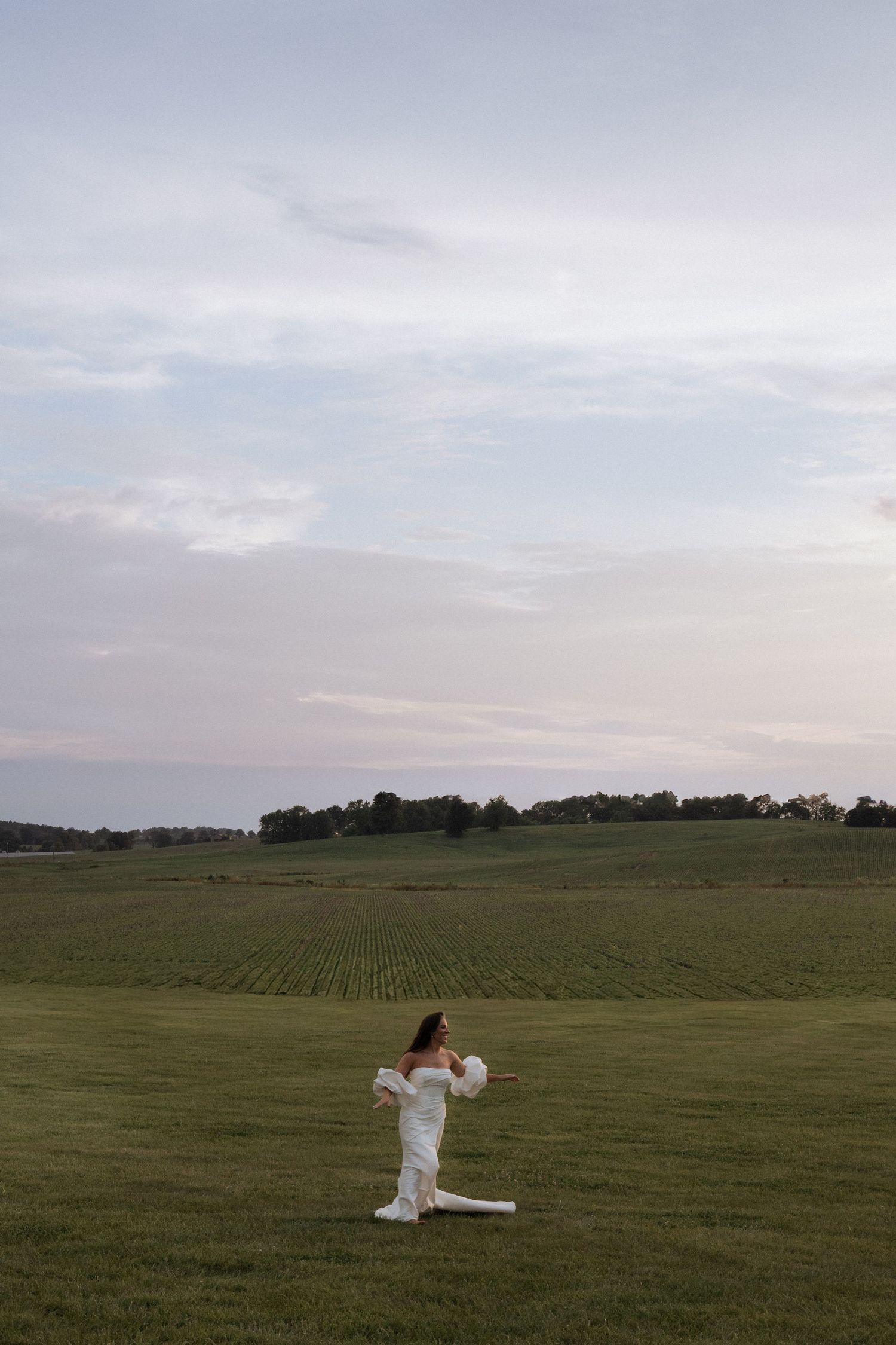 A woman in a white dress stands gracefully in a vast green field under a cloudy sky at dusk.