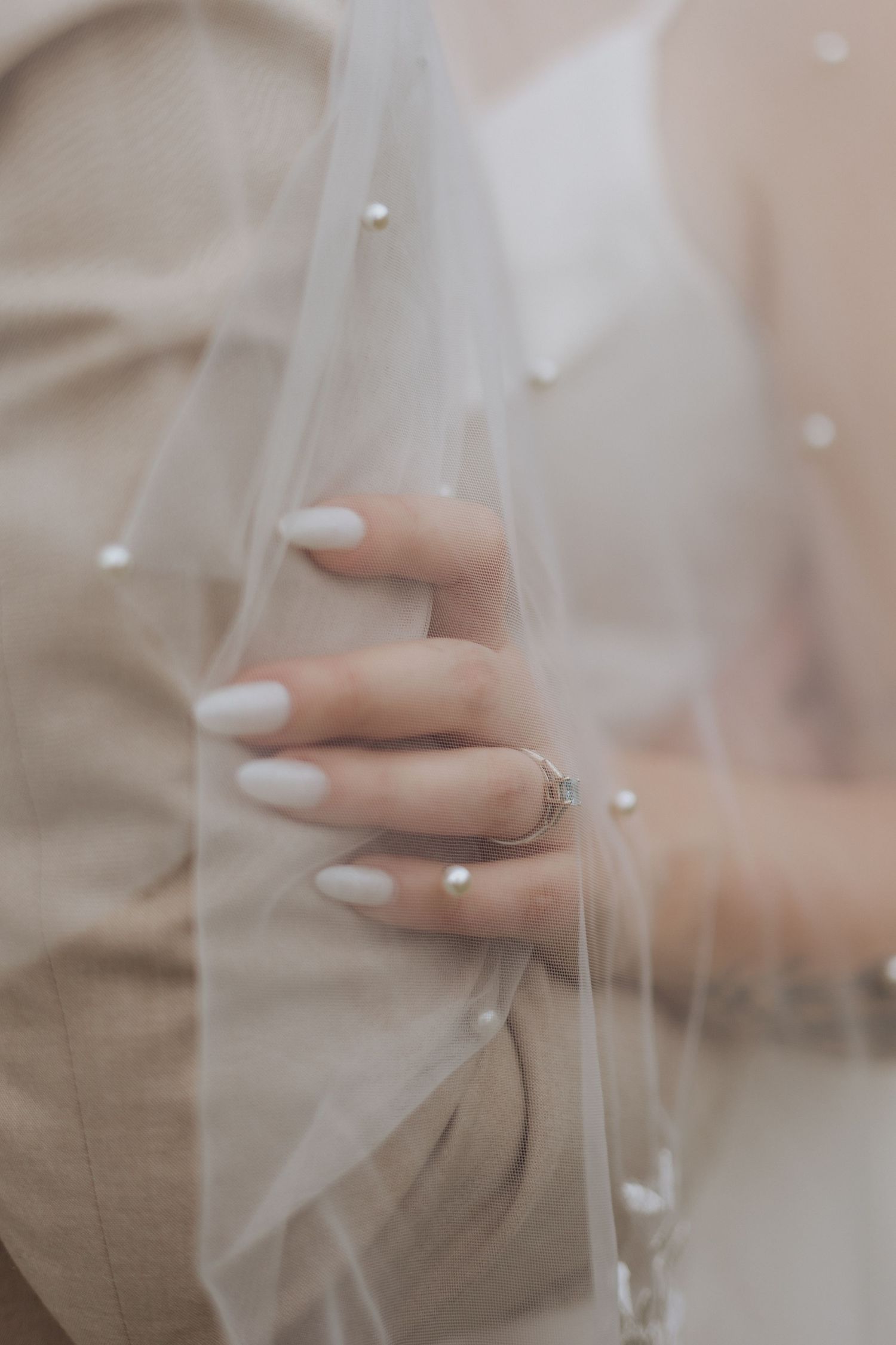 Close-up view of a delicate engagement ring on a hand with white nail polish against a soft, ethereal veil background.