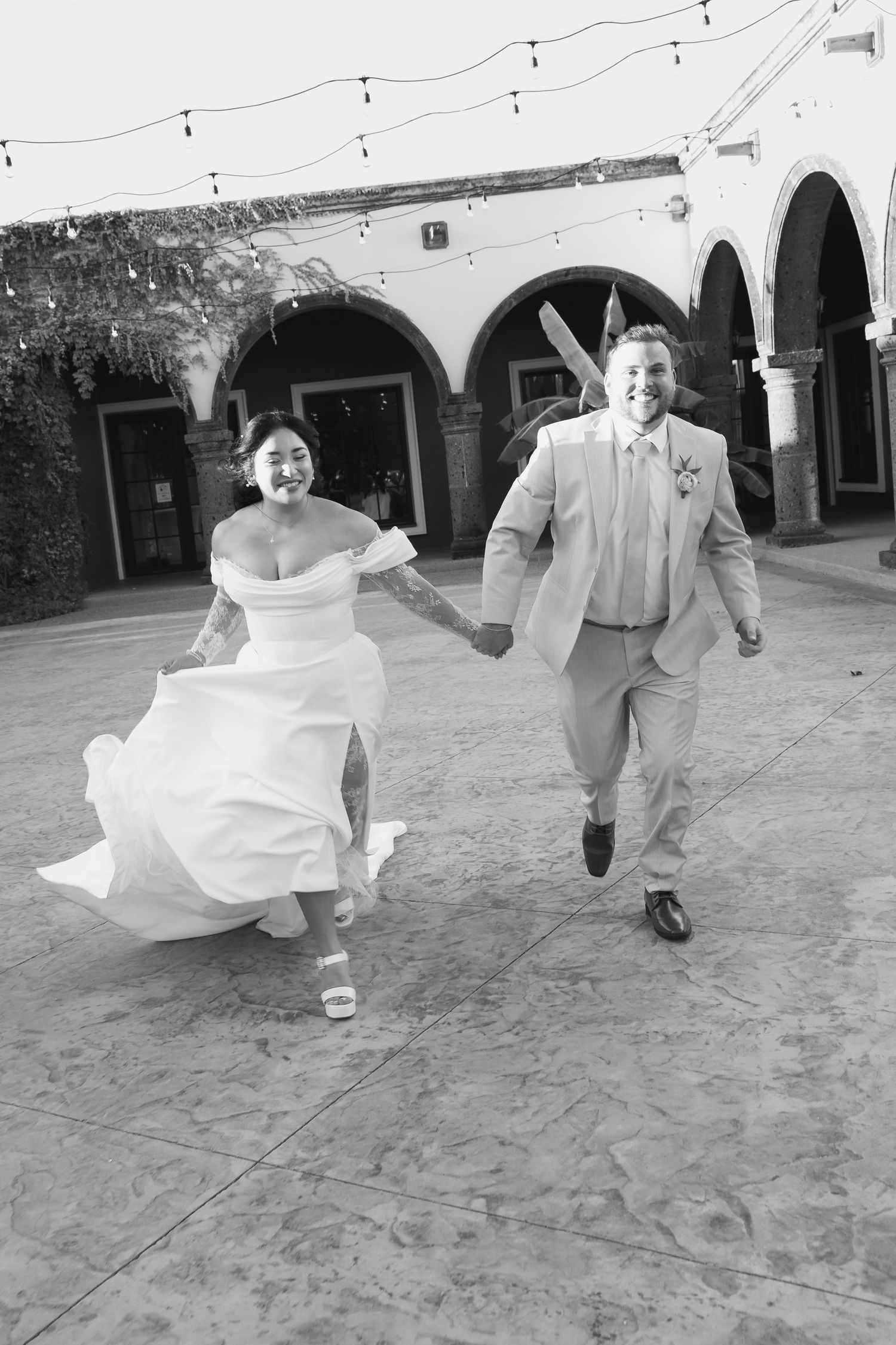 A joyful couple runs together during their wedding celebration under Spanish-style arches with string lights overhead.