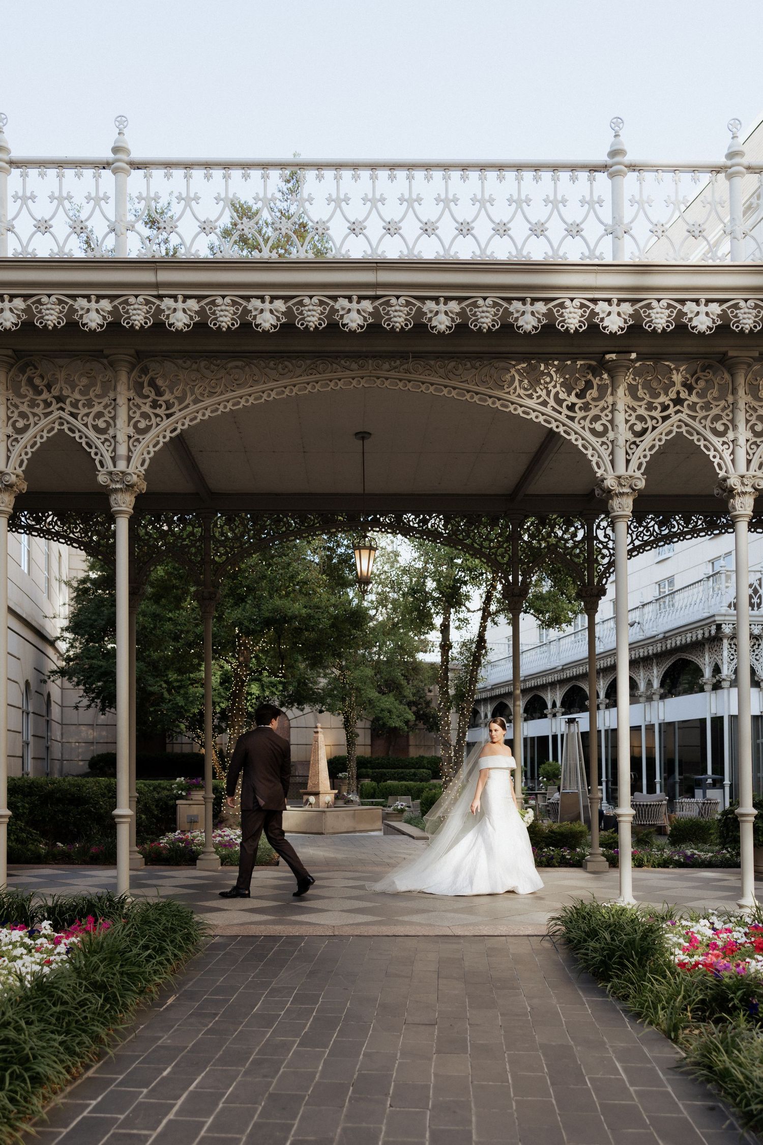 Bride and groom walk under ornate white iron gazebo in lush garden courtyard on their wedding day.