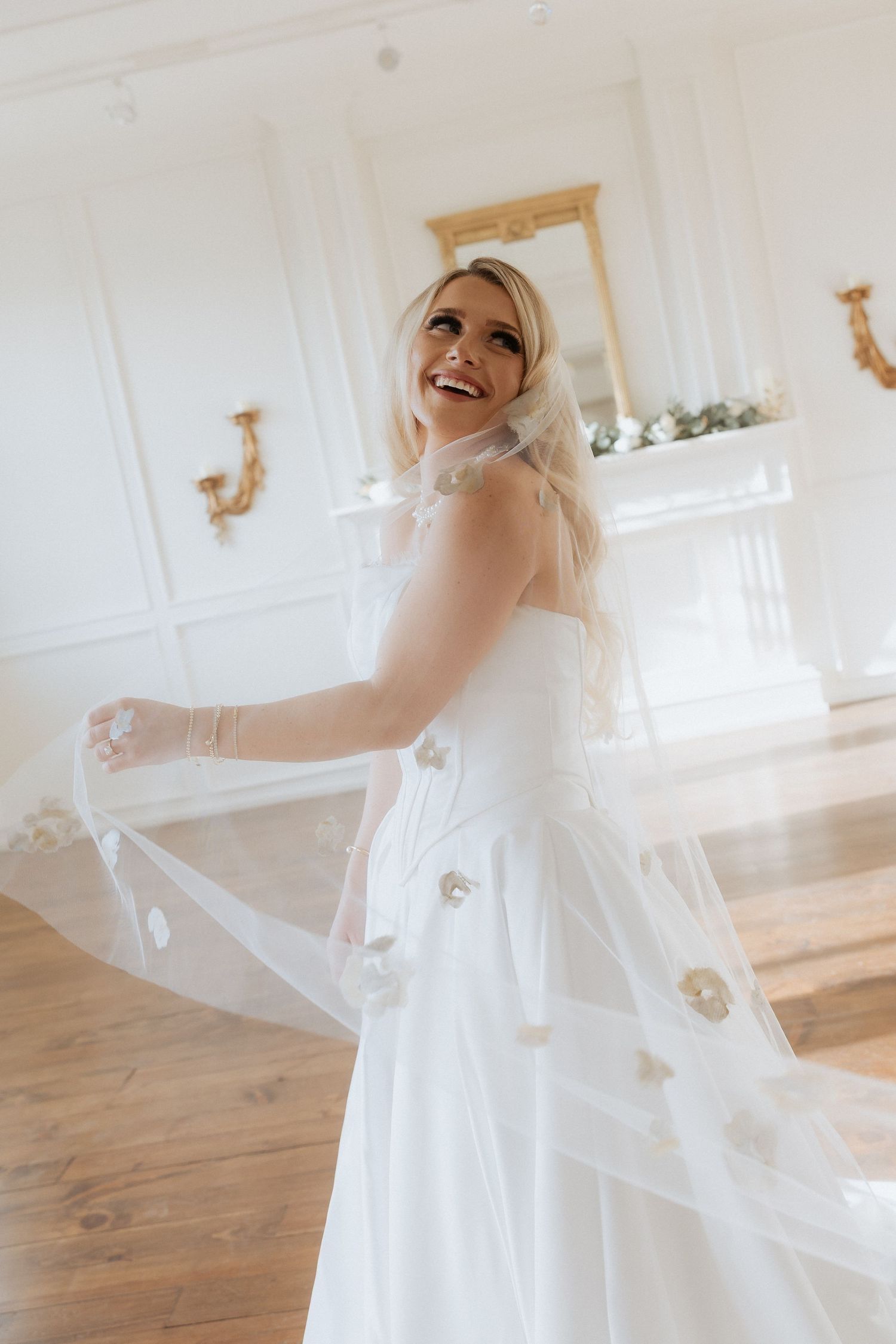 A joyful bride in a flowing white wedding dress twirls and dances in a bright, sunlit room with wooden floors.