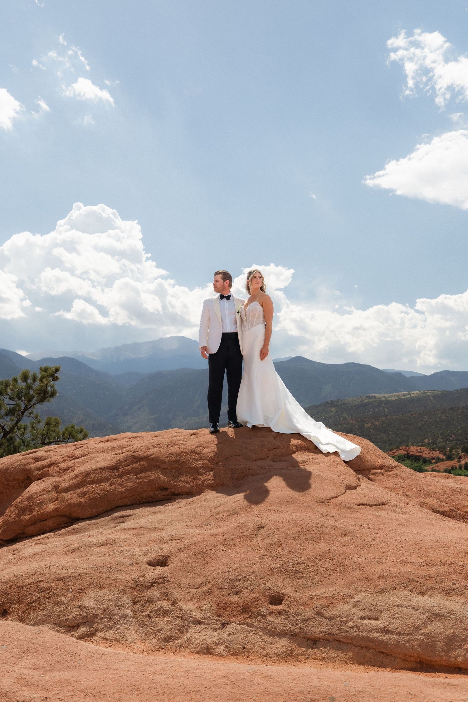 Bride and groom stand atop red rock formation at Garden of the Gods with mountain backdrop and blue sky.