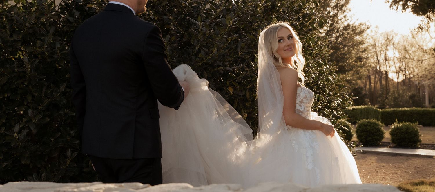 Bride in elegant white ball gown and veil walks with groom outdoors at a garden wedding venue.