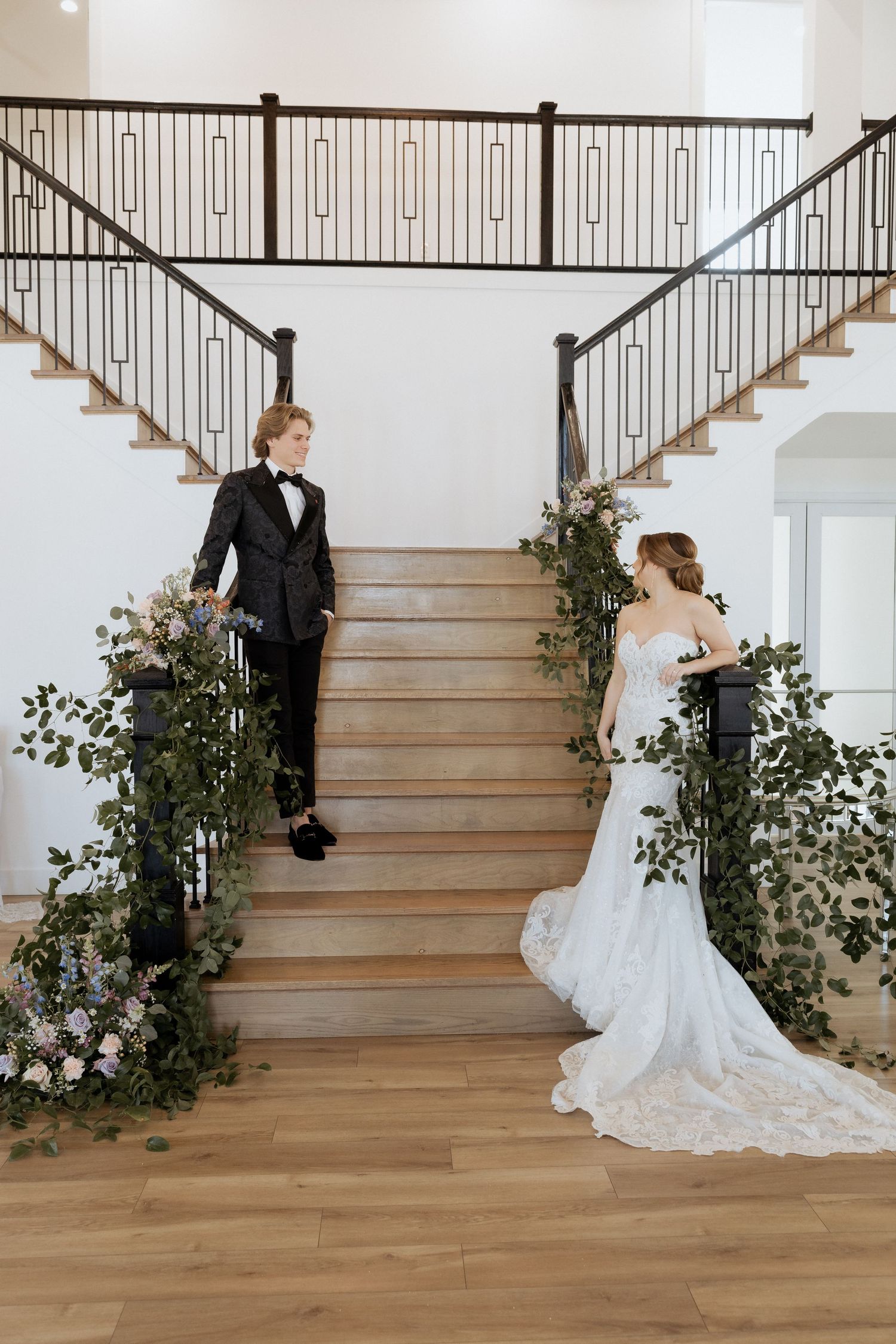 Elegant couple poses on a wooden staircase adorned with greenery and floral arrangements in a bright interior space.