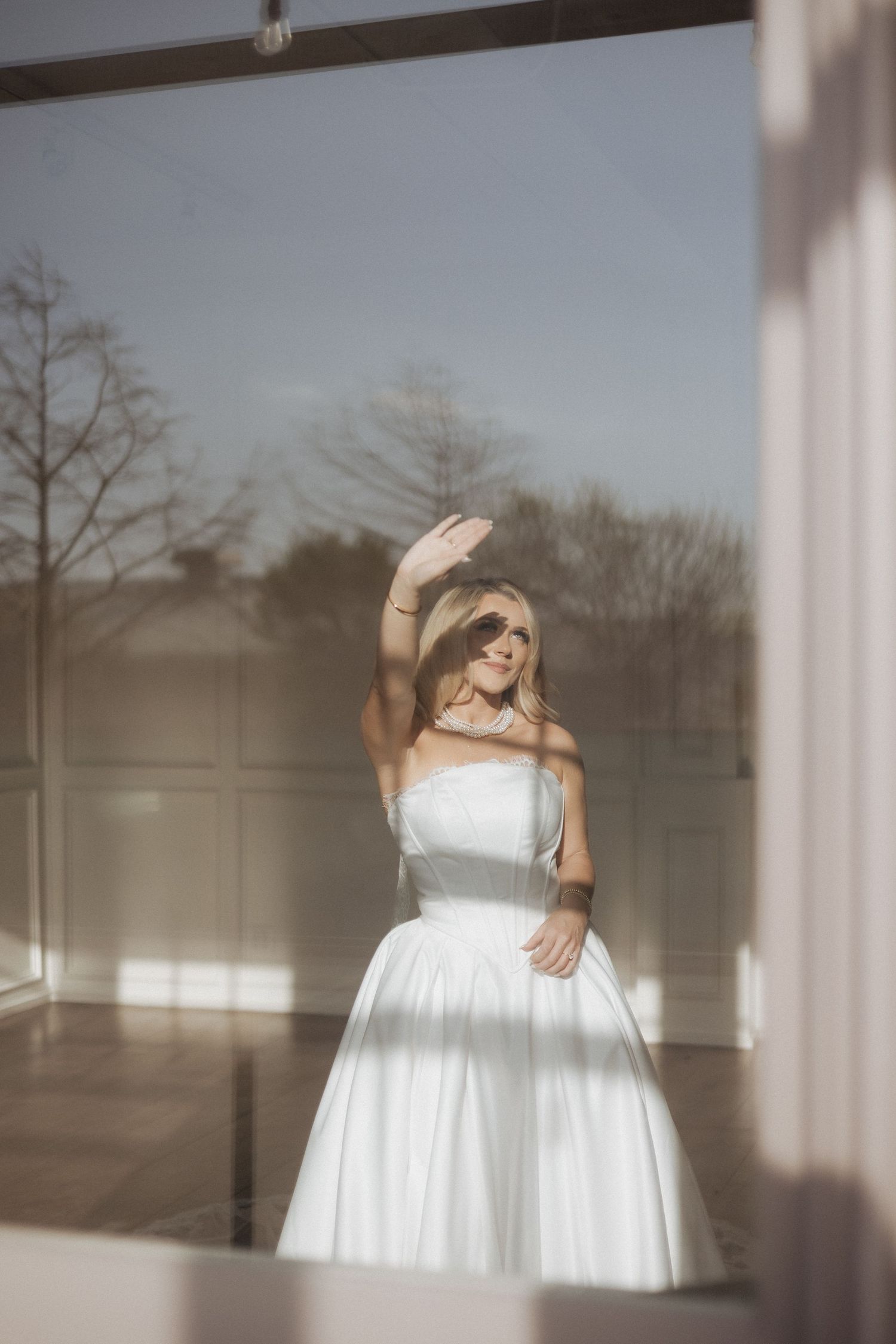 A bride in a classic white strapless ball gown waves through a window, creating an ethereal silhouette against natural light.