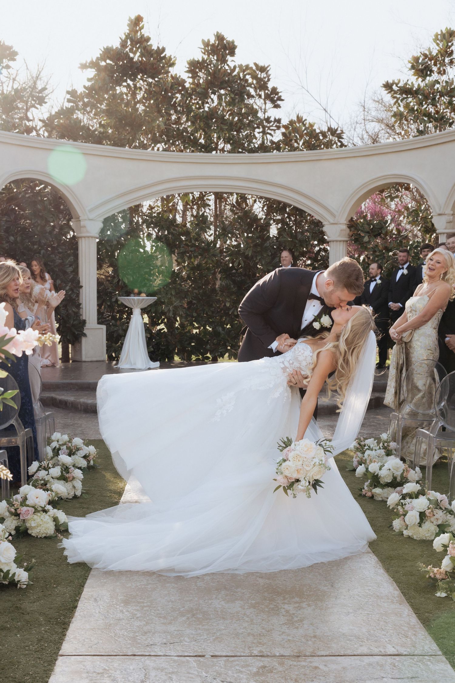 Groom dips bride for a kiss at outdoor wedding ceremony with white floral arrangements and elegant arched backdrop.