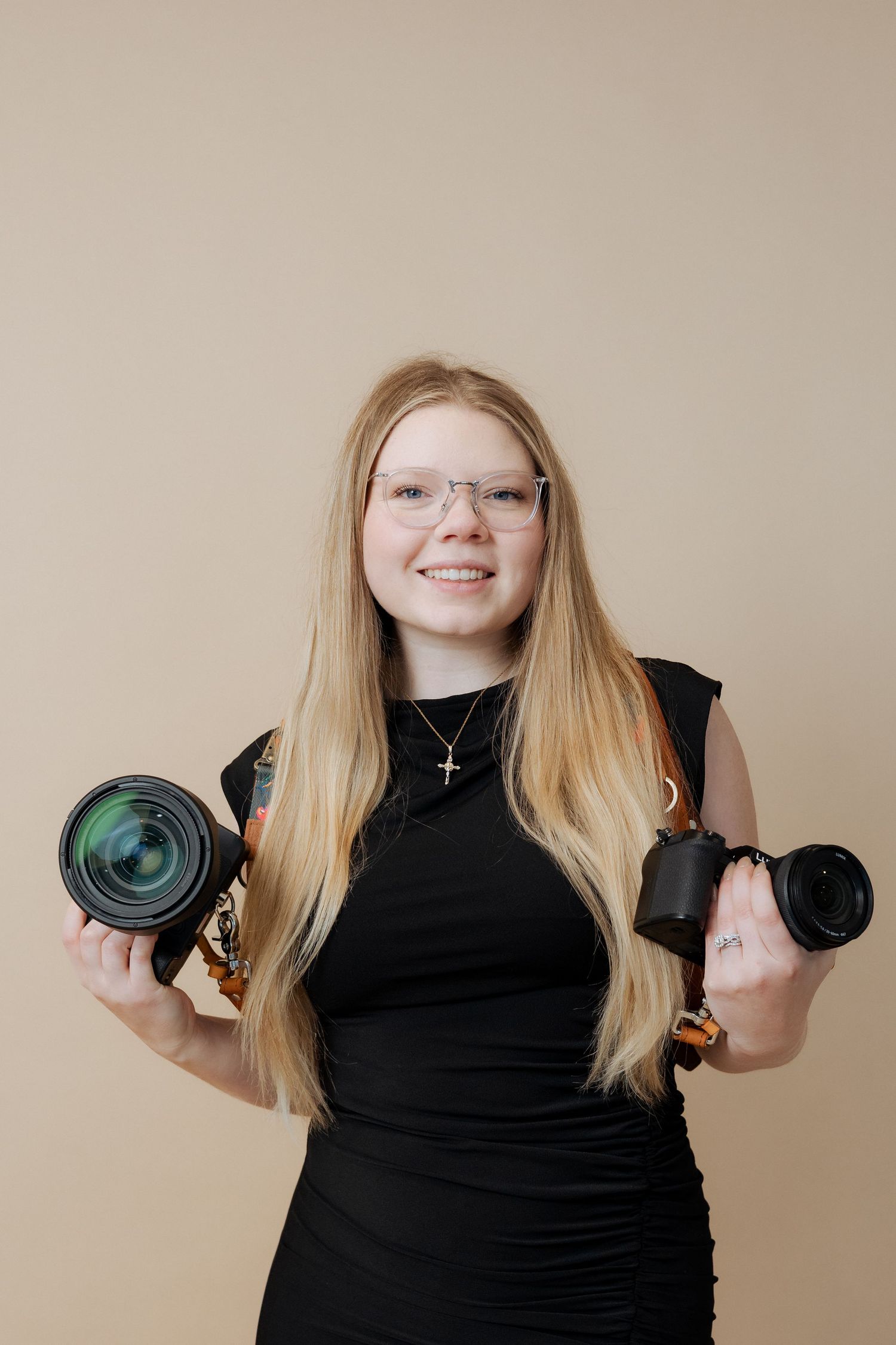 A photographer in a black dress holds two different camera lenses against a beige background.