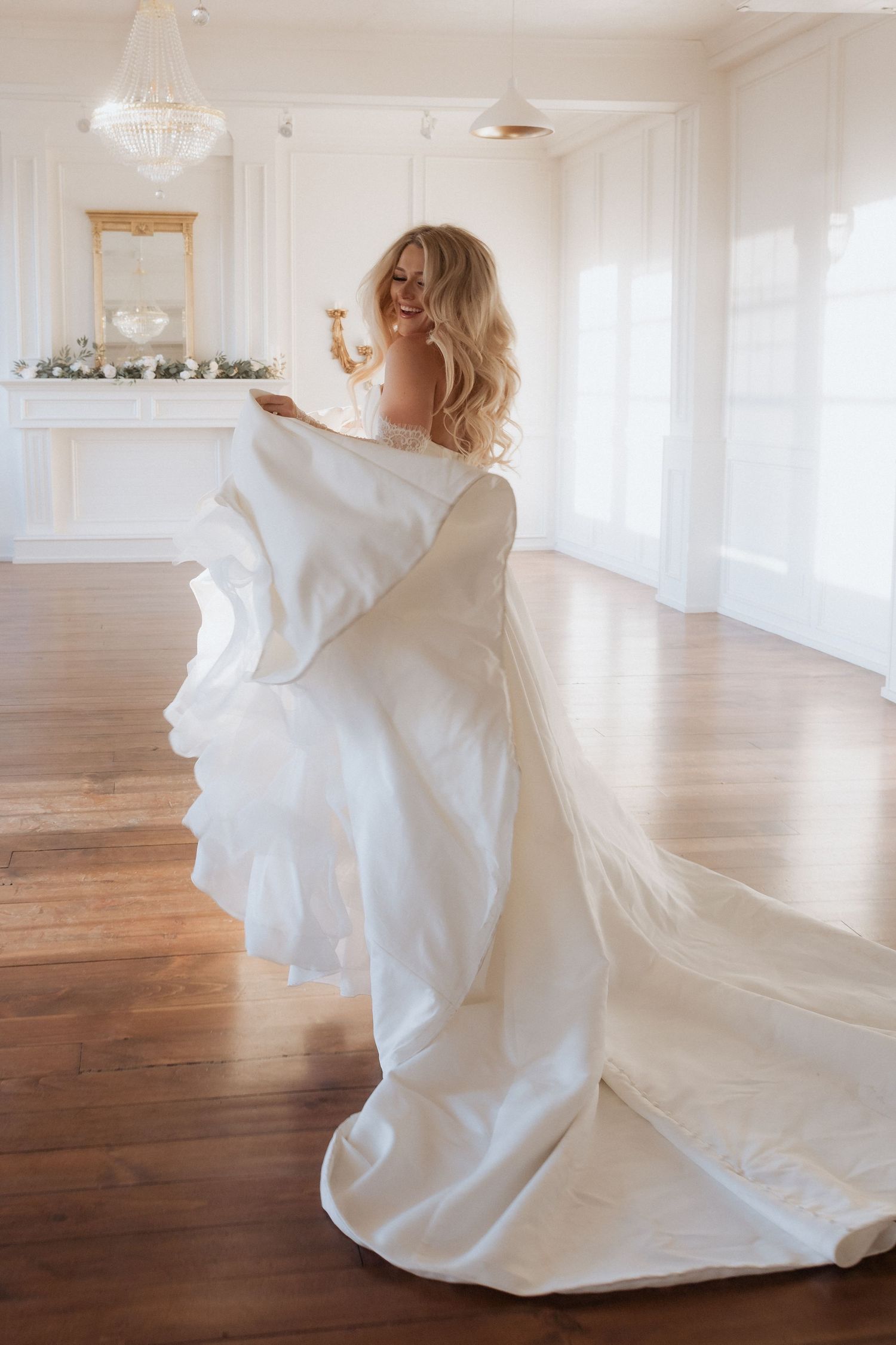 A bride in a flowing white wedding gown twirls gracefully across a bright sunlit room with hardwood floors.