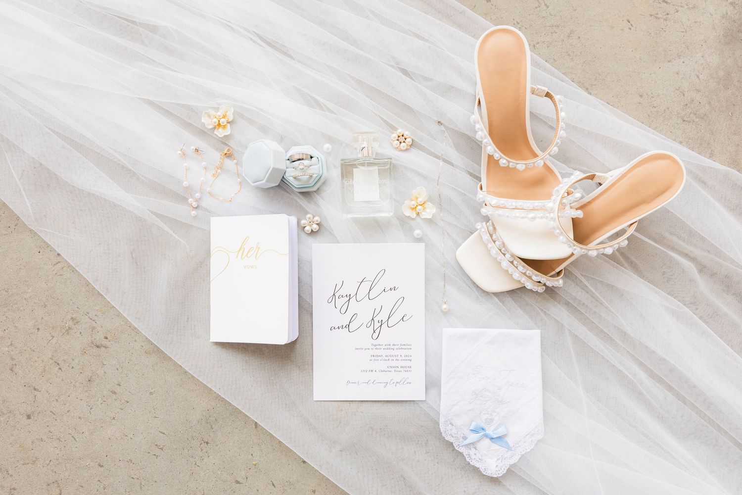 Elegant wedding flatlay with white invitations, delicate jewelry, and strappy high heel sandals on light fabric background.