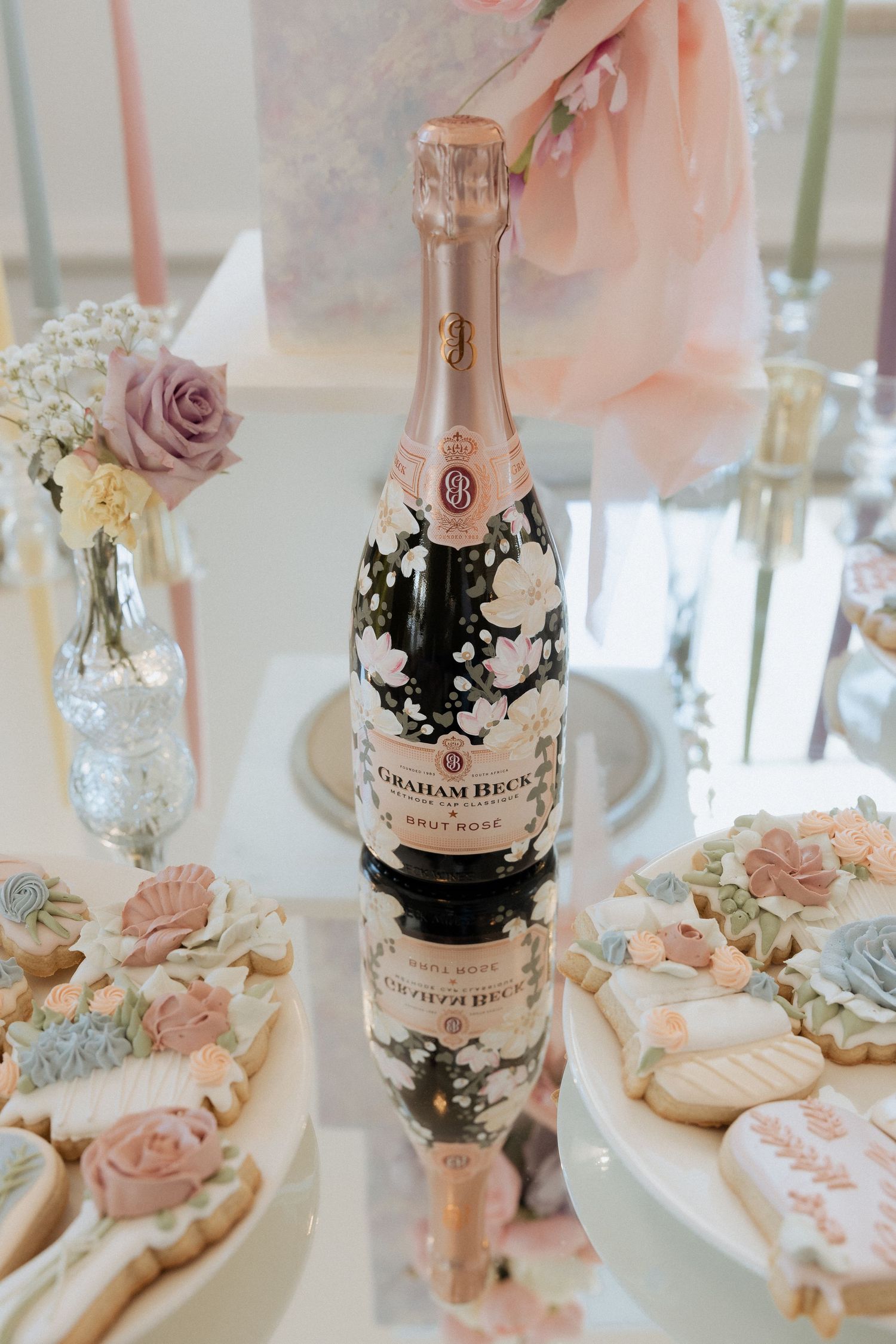 A bottle of pink Graham Beck rosé champagne sits on a mirrored surface with delicate floral decorations.
