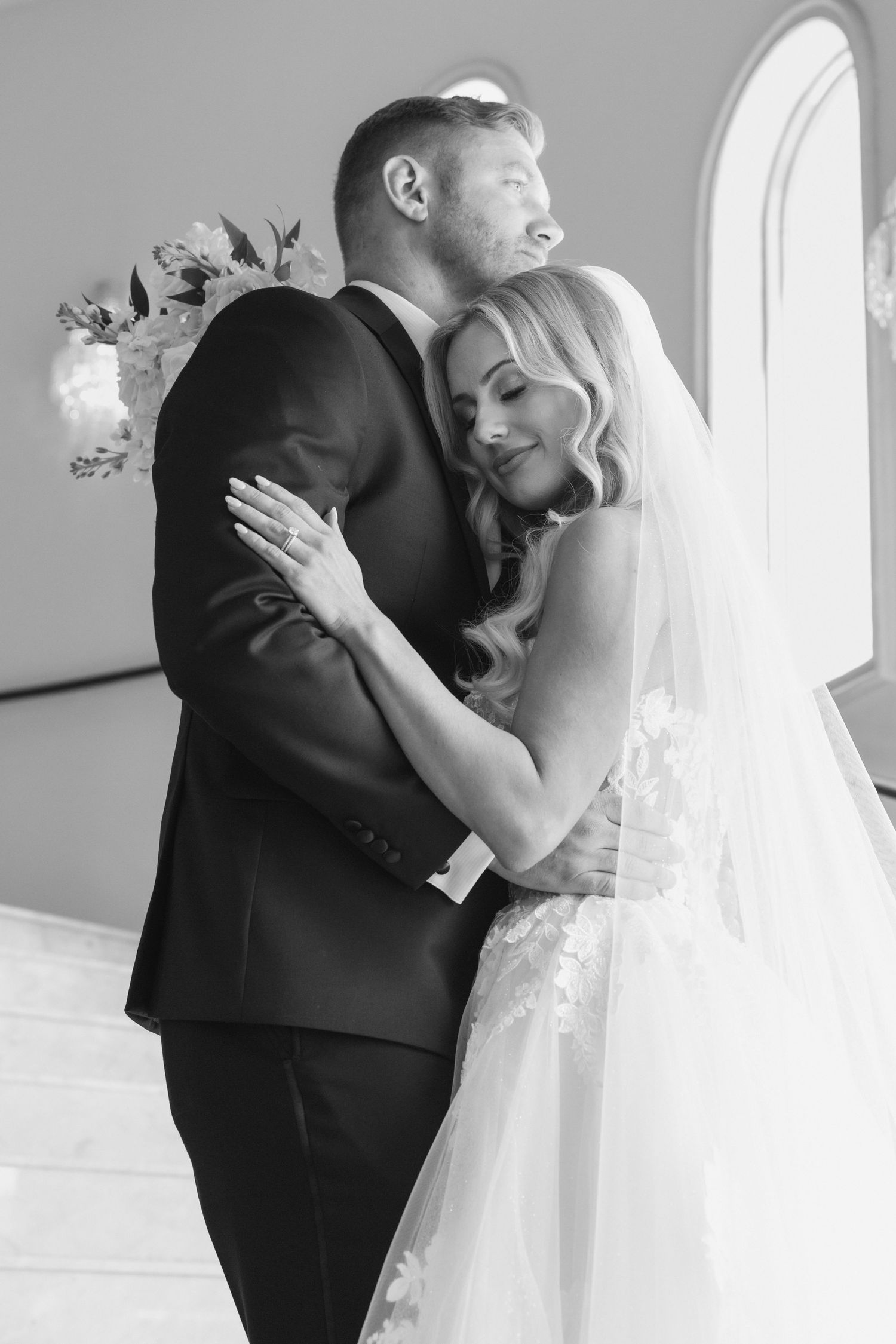 Black and white wedding photo of bride and groom embracing tenderly in a bright chapel setting.