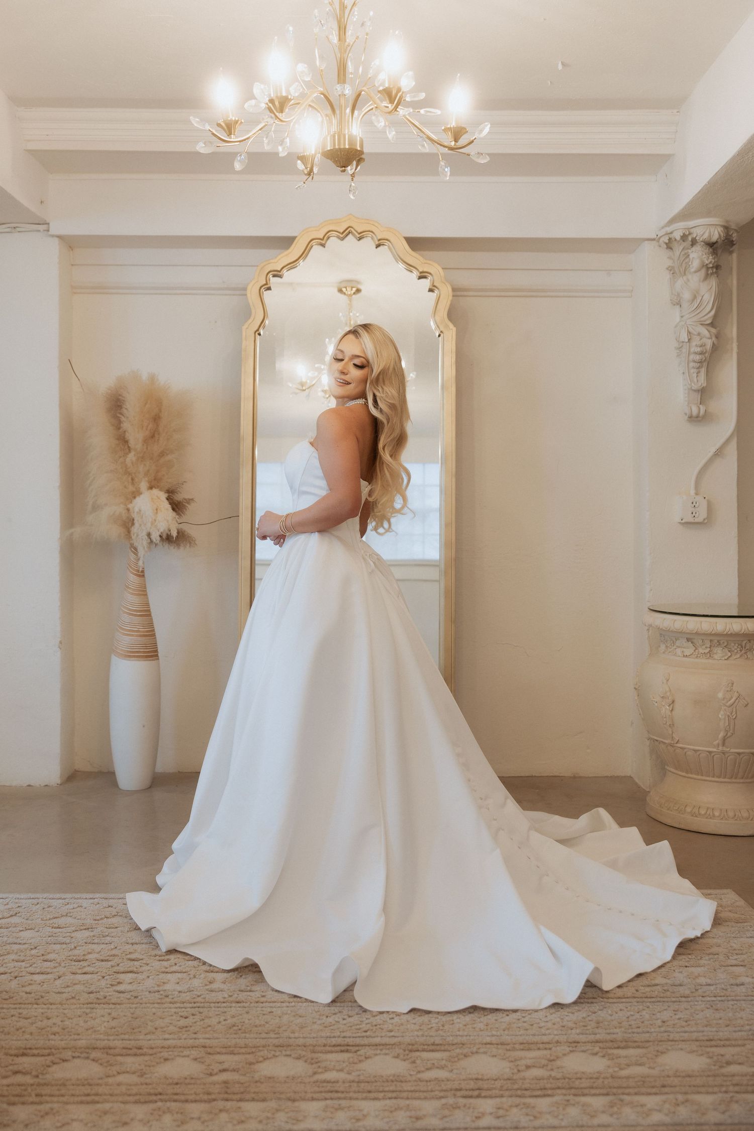 A stunning ballgown wedding dress with a flowing train on display in an elegant room with ornate gold mirror and chandelier.