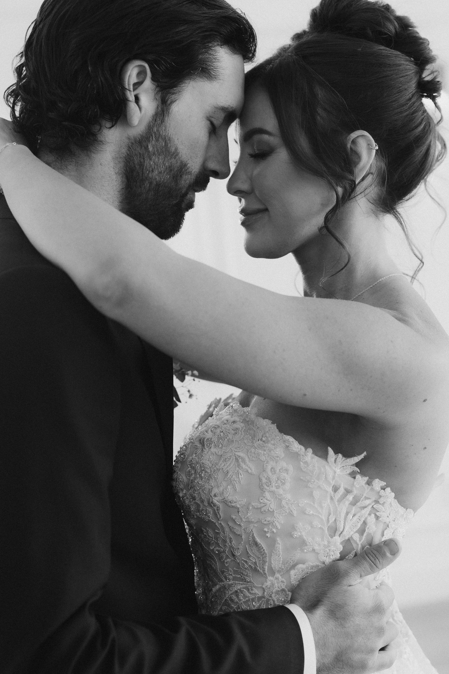 A romantic black and white wedding photograph showing an intimate moment between a bride in lace and her bearded groom.