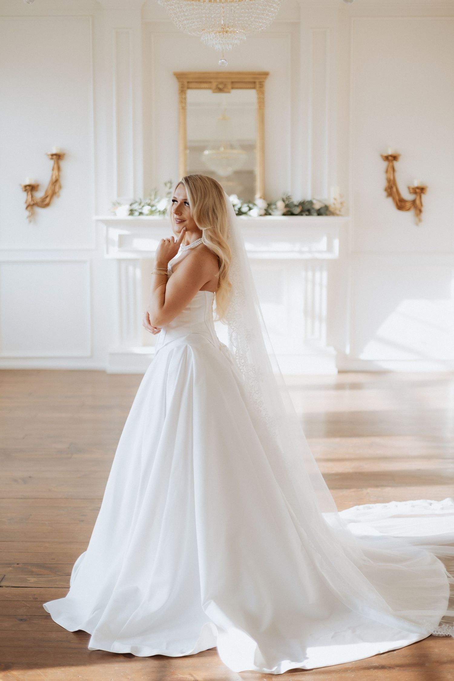 A classic white wedding gown with flowing train is showcased in an elegant room with ornate wall sconces and wooden floors.