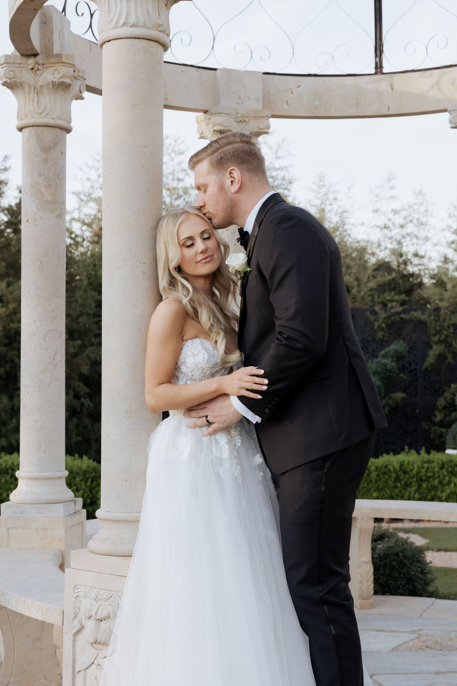 A bride and groom share a romantic moment under an elegant stone gazebo surrounded by lush greenery on their wedding day.