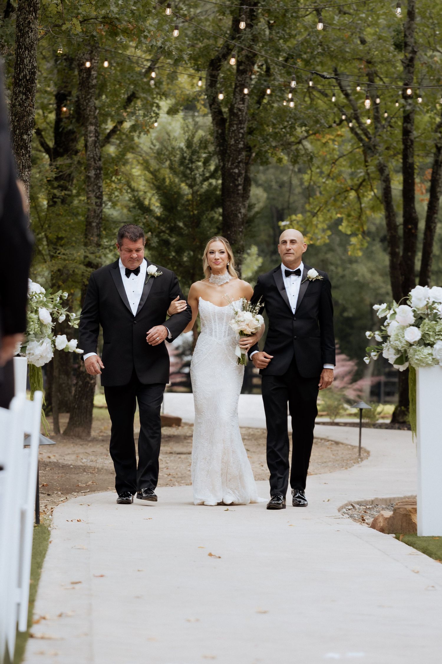 Wedding party members in black tuxedos and white dresses walk together on a tree-lined path outdoors.