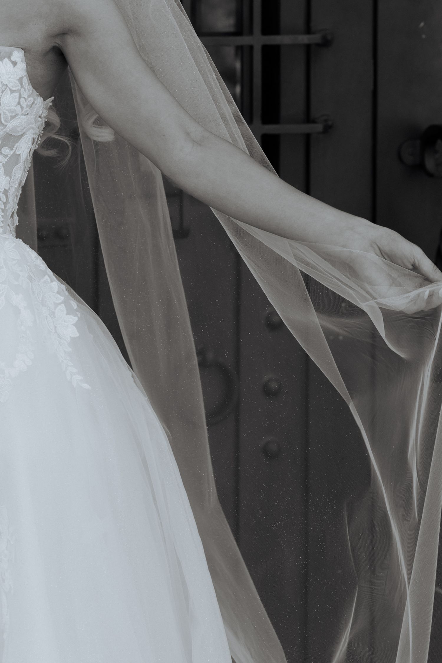 Bride in white lace gown holding flowing veil near ornate dark metal door, black and white wedding photo.