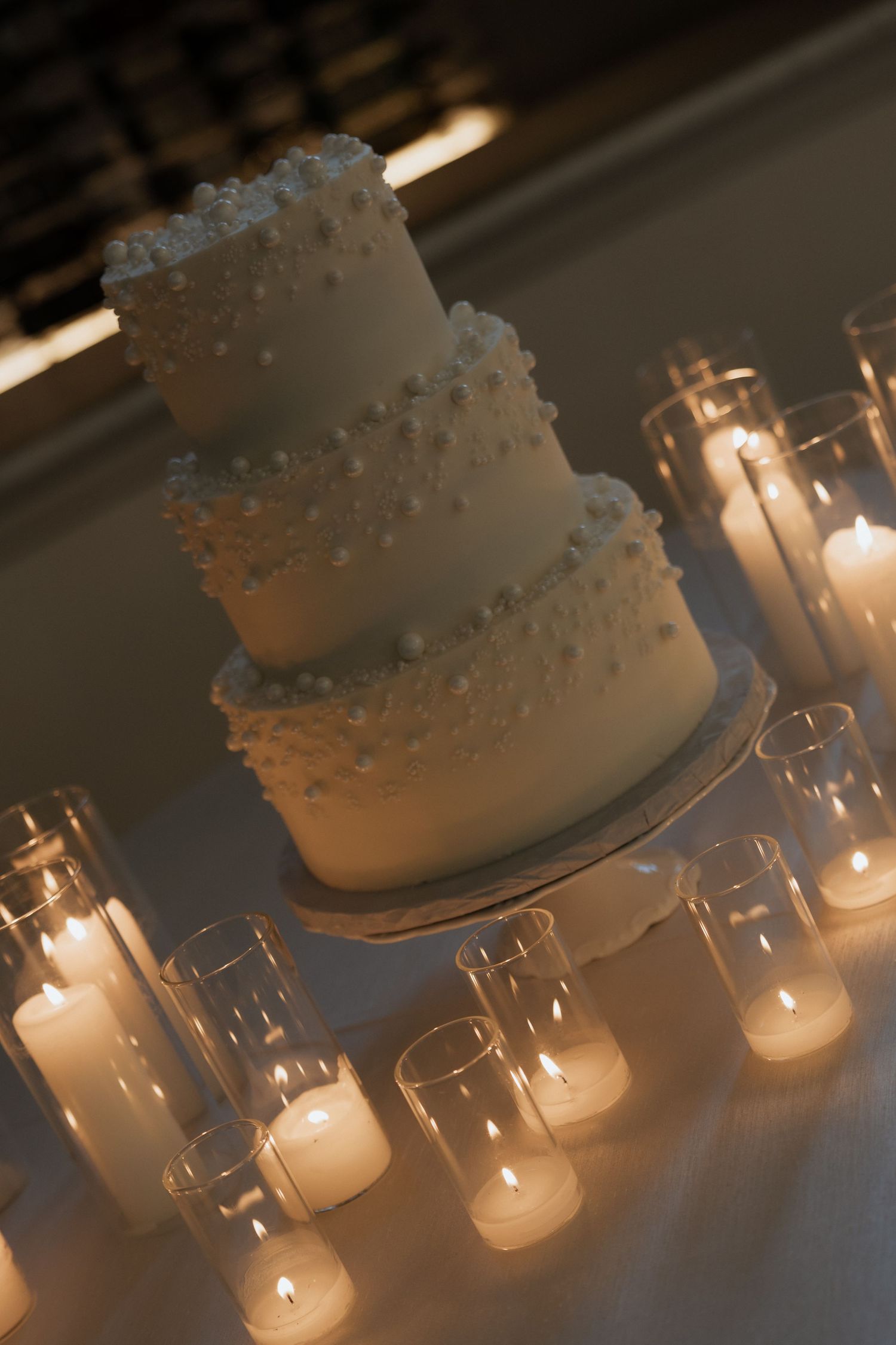 Three-tier white wedding cake adorned with pearl details, surrounded by glowing pillar candles in glass holders.