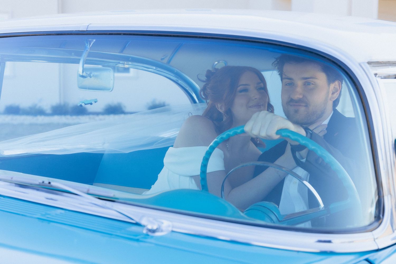 Two people sit in a classic light blue vintage car on a sunny day through the windshield view.