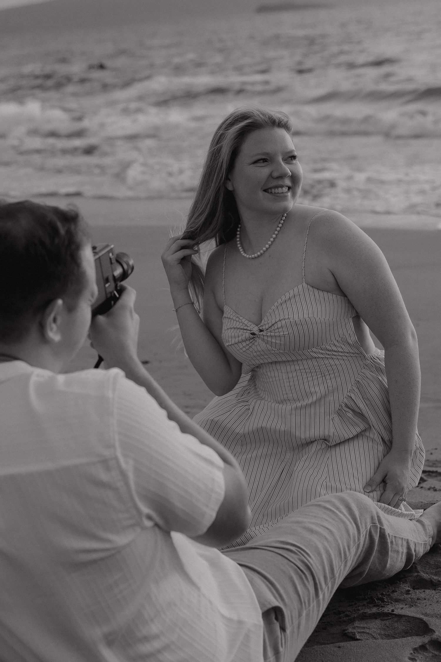 Black and white beach photo of a woman in striped dress and pearls smiling while a man photographs her by the ocean.