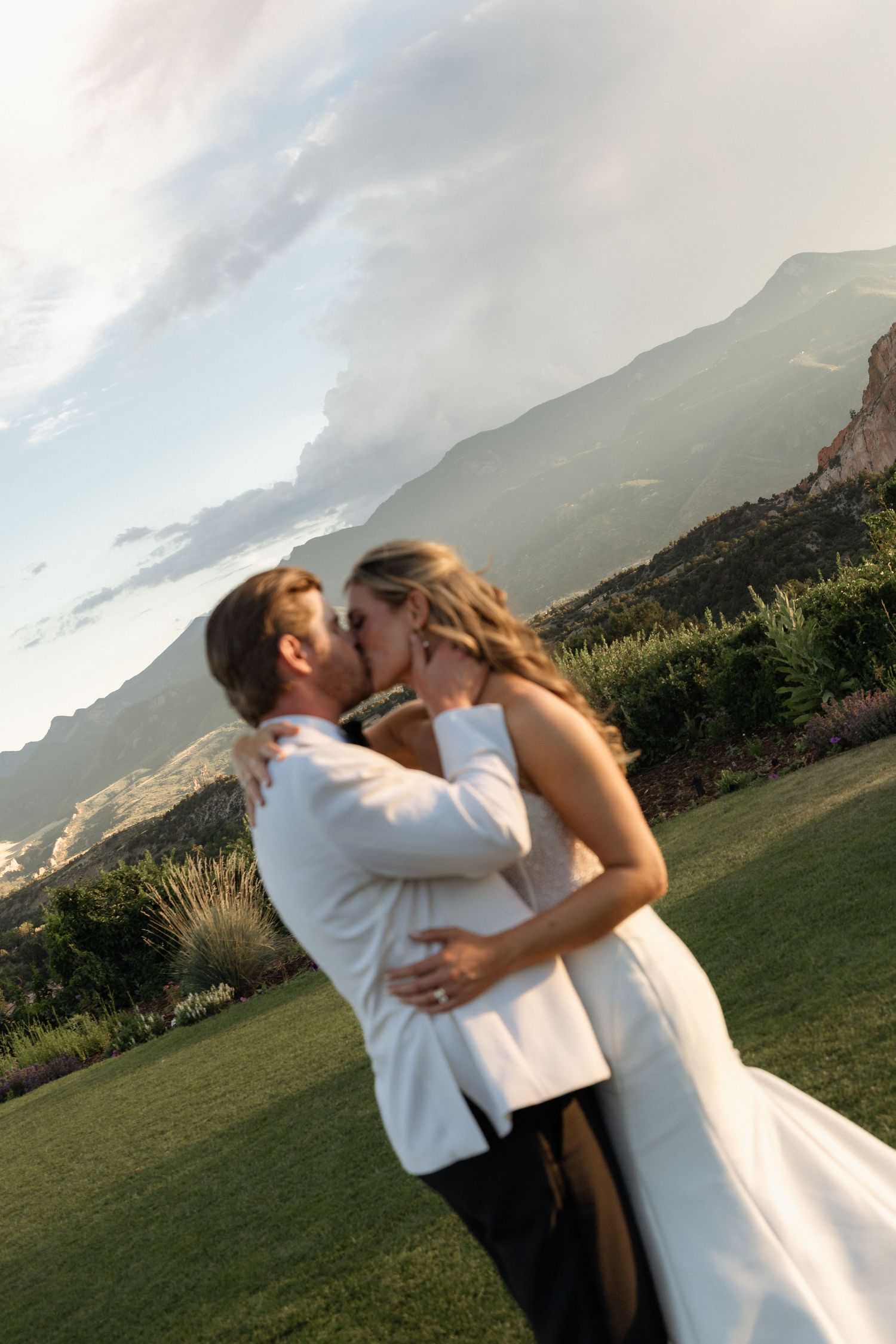 Bride and groom share a romantic kiss on a green lawn with stunning mountain backdrop during outdoor wedding ceremony.