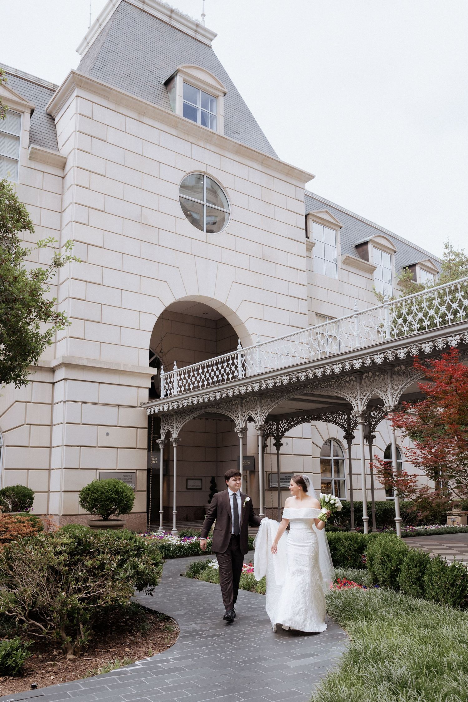 Bride and groom walk hand in hand outside an elegant historic venue with ornate iron balcony and lush gardens.