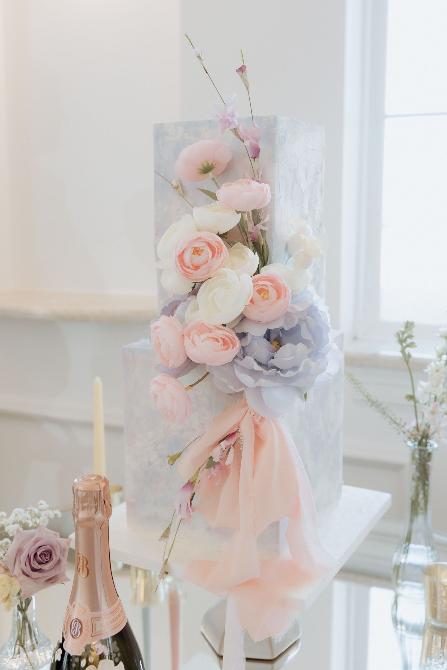 Elegant wedding cake decorated with delicate pink and gray sugar flowers and tulle draped alongside a champagne bottle.