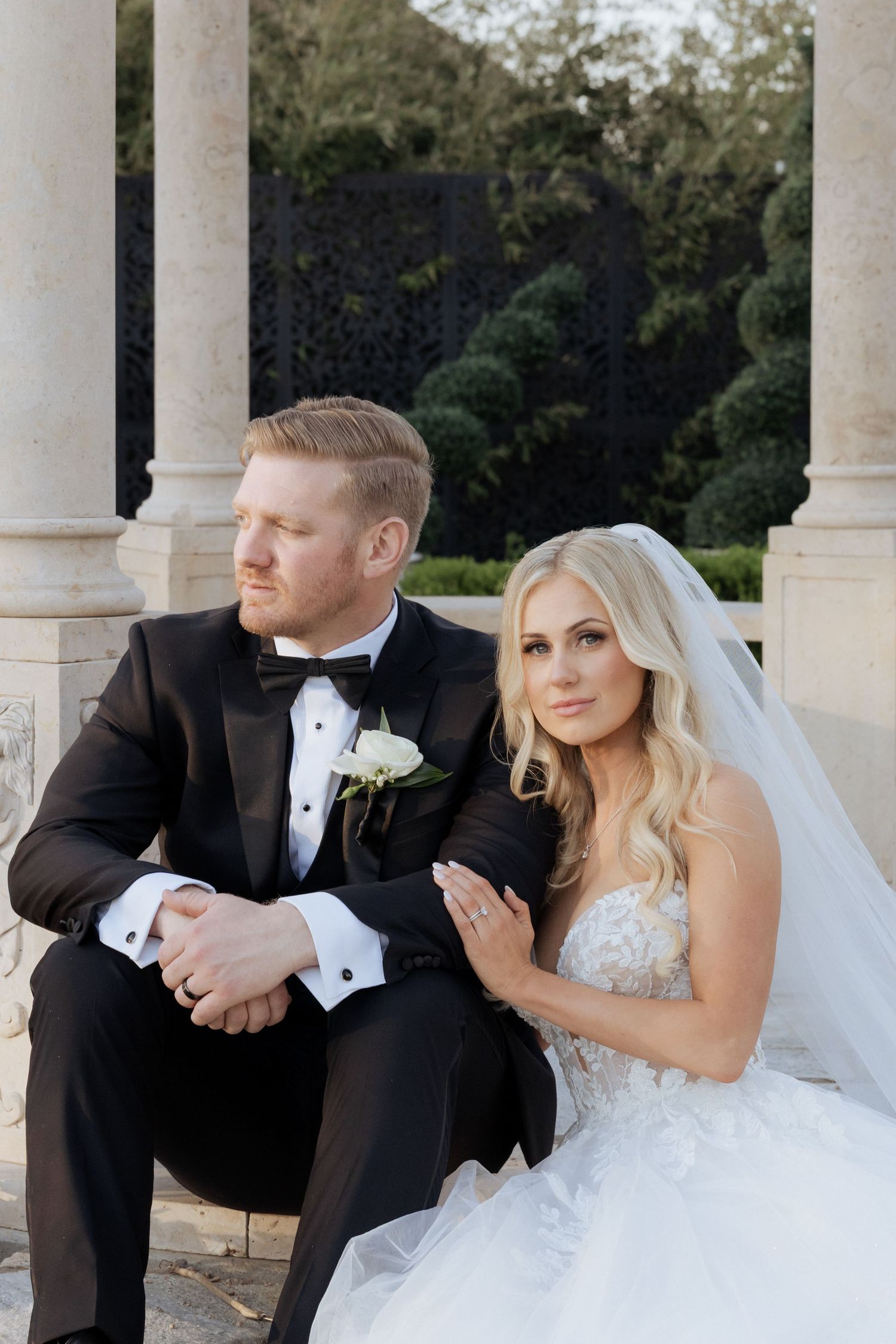 A bride in a white lace gown and veil sits beside a groom in a black tuxedo among elegant stone columns.