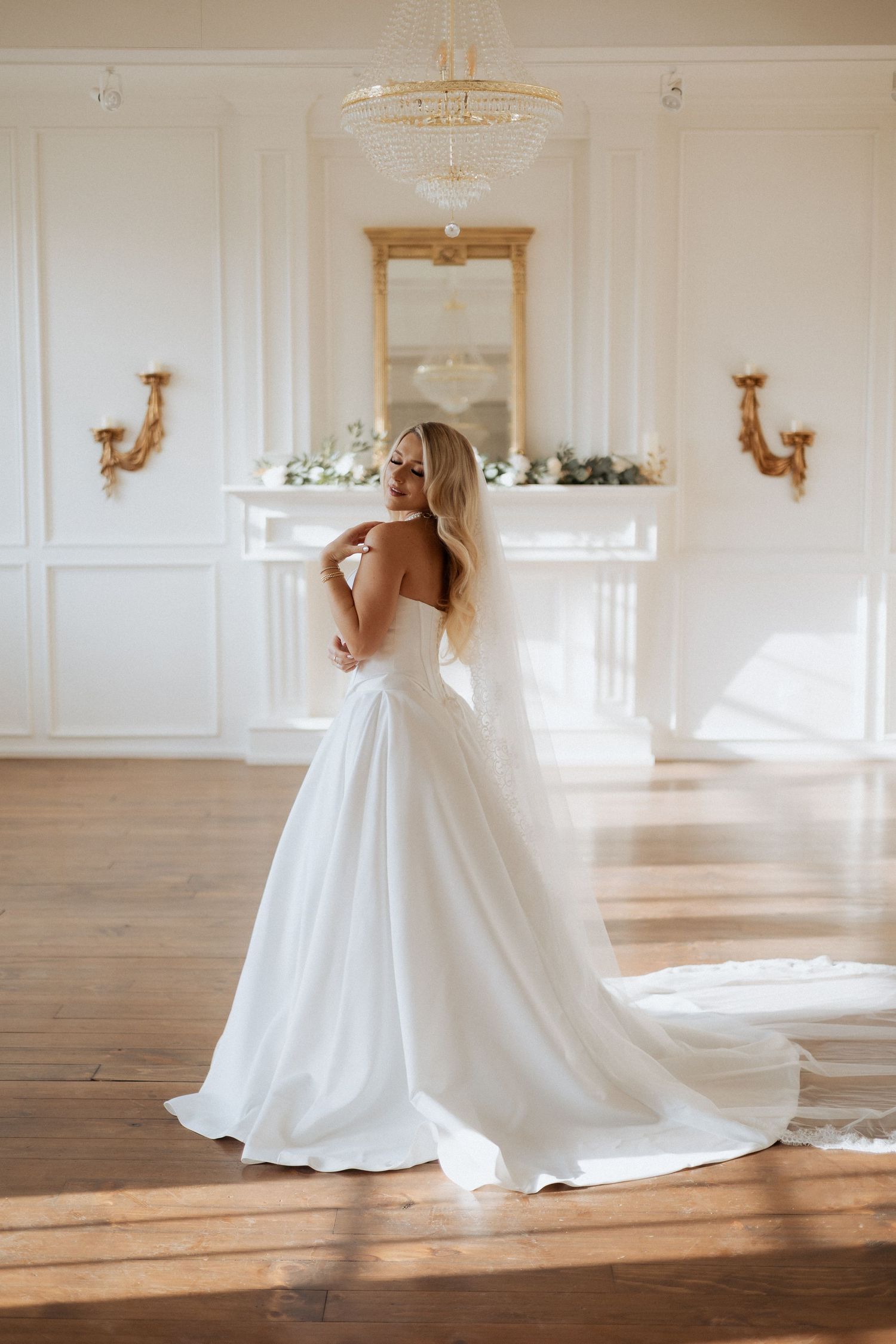Elegant white wedding gown with strapless bodice and flowing train in bright sunlit room with ornate wall sconces.